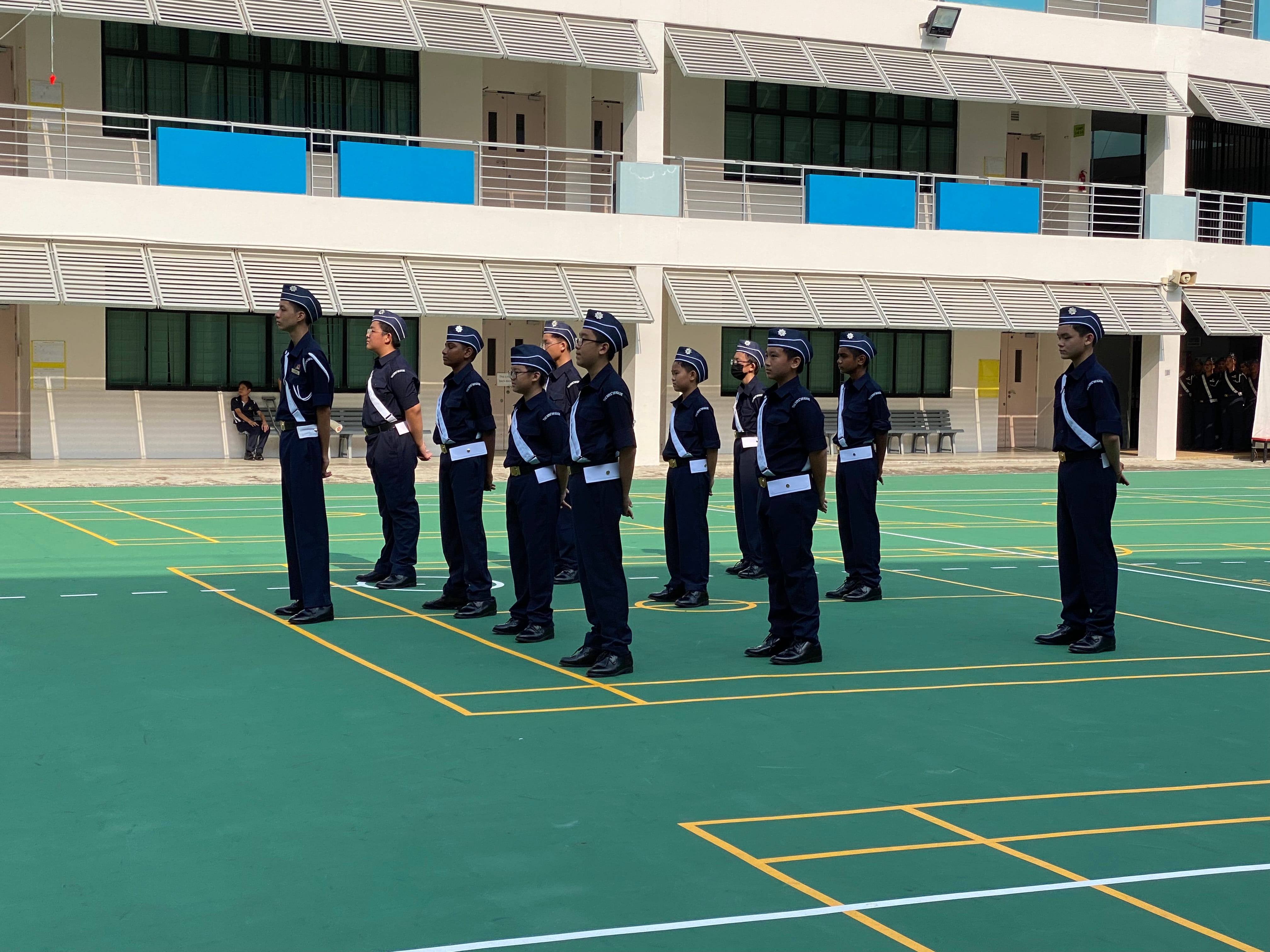 Cadets in navy uniforms and caps stand at attention in two rows on a green outdoor court beside a white building.