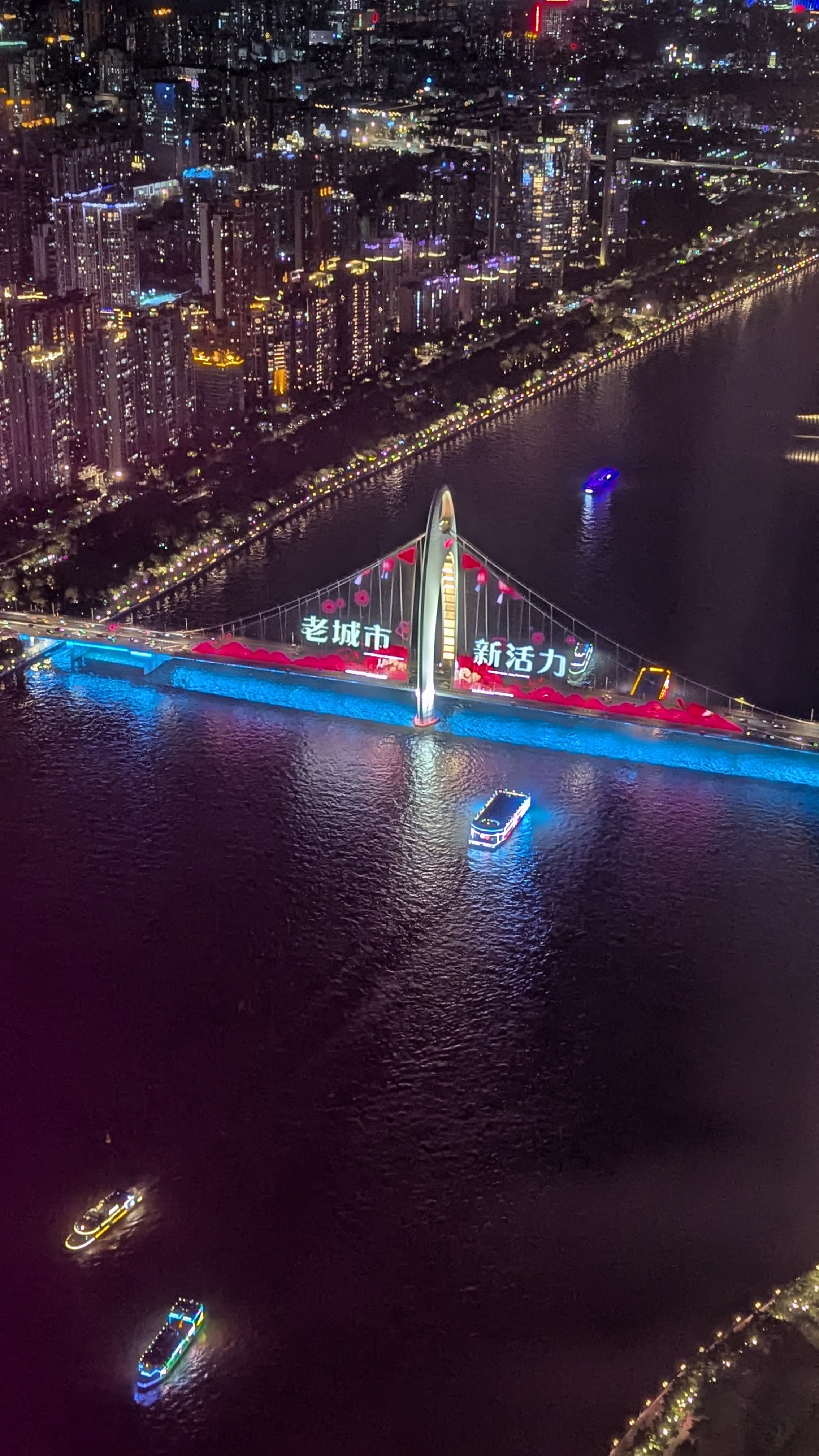 Aerial night view of a illuminated suspension bridge with Chinese text over a dark river, city lights in background.