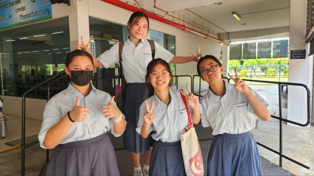 Four female students in light blue uniforms pose with peace signs and thumbs up outside a school building.