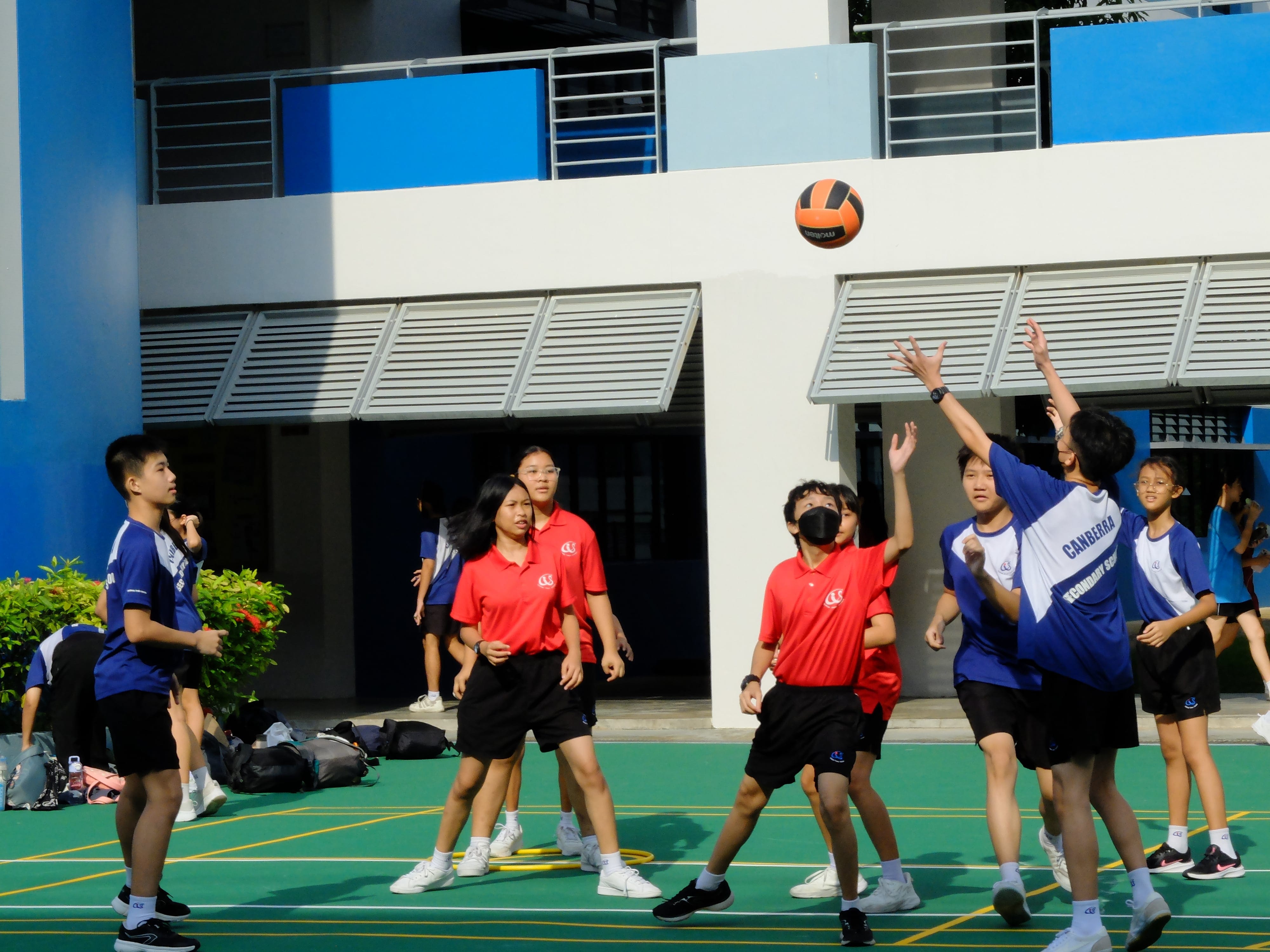 Students in red and blue uniforms play volleyball on an outdoor court beside a blue and white school building.