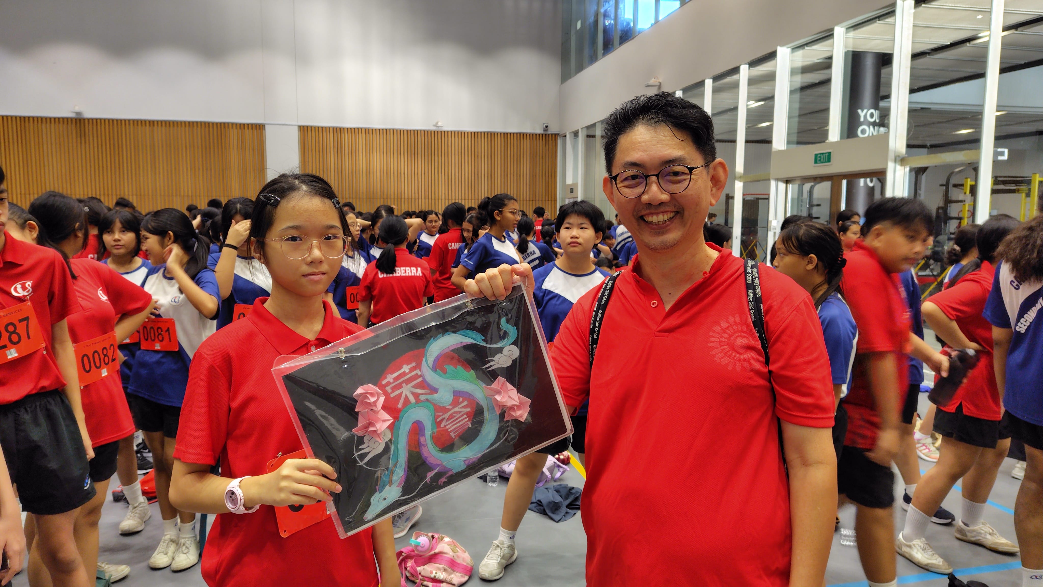 A girl and a man in red shirts hold a framed dragon artwork inside a school hall filled with students.
