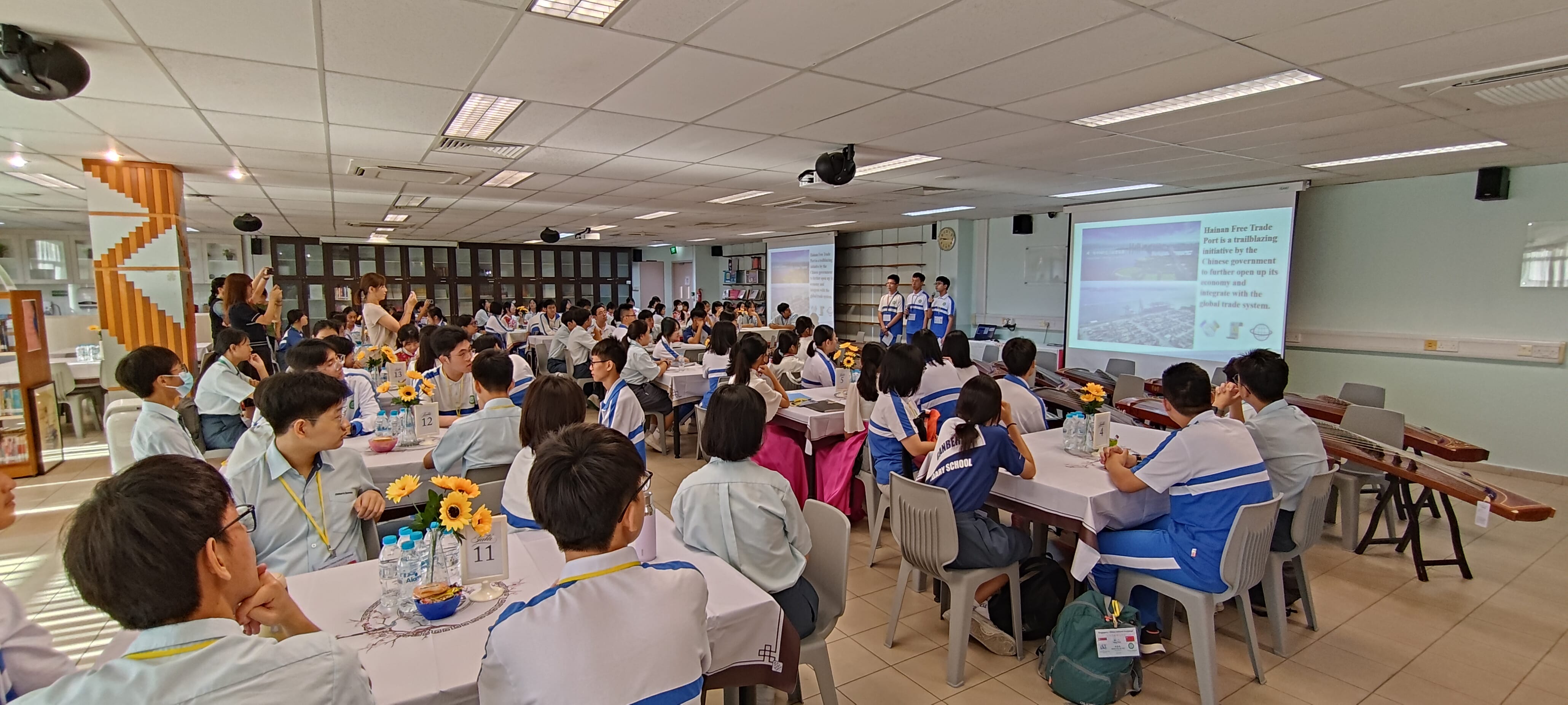 Students in uniforms sit at tables with sunflowers watching a presentation on a projected screen in a school hall.