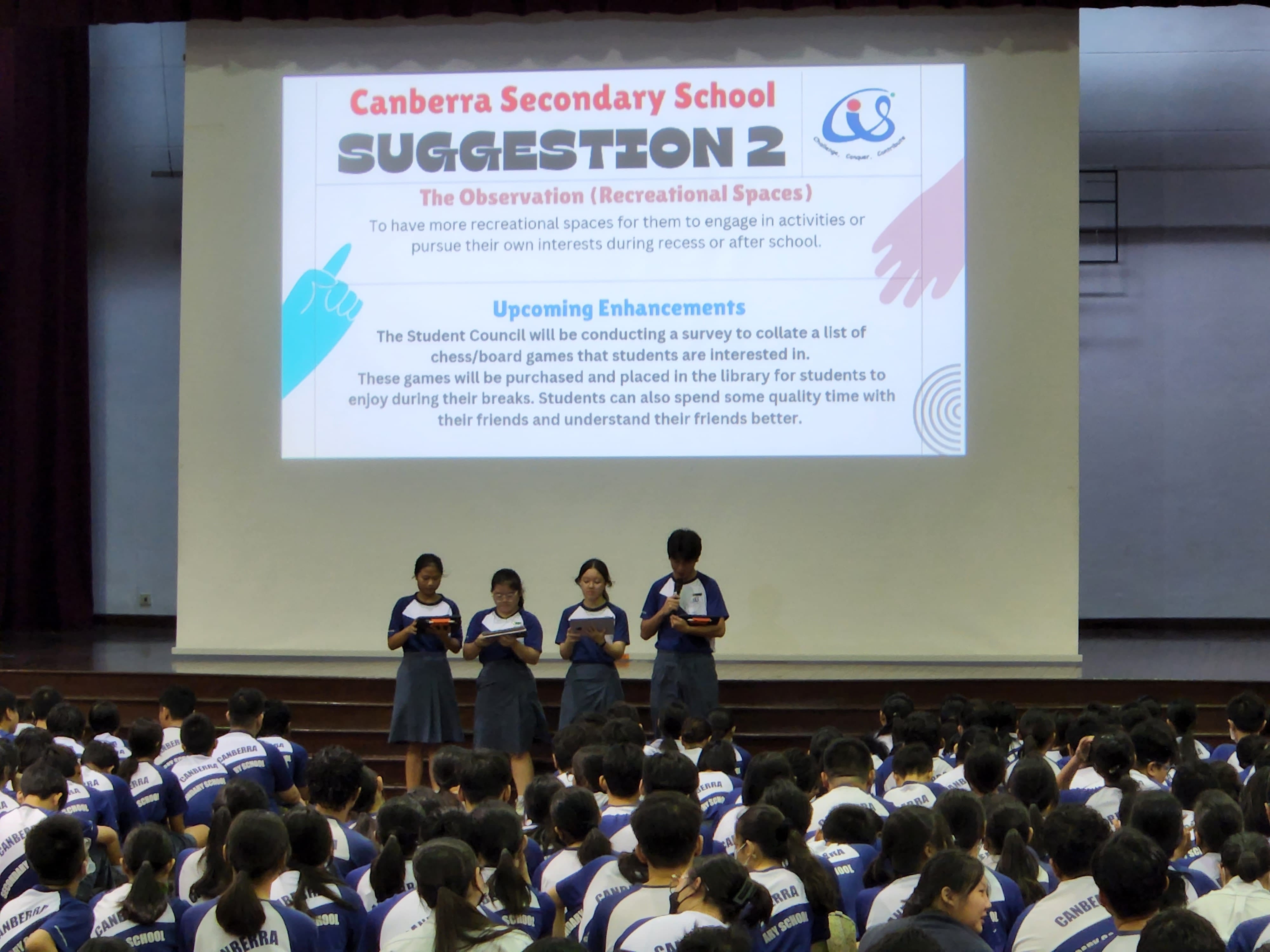 Four students address the school in the school hall, with the words "Suggestion 2' on a large screen behind them