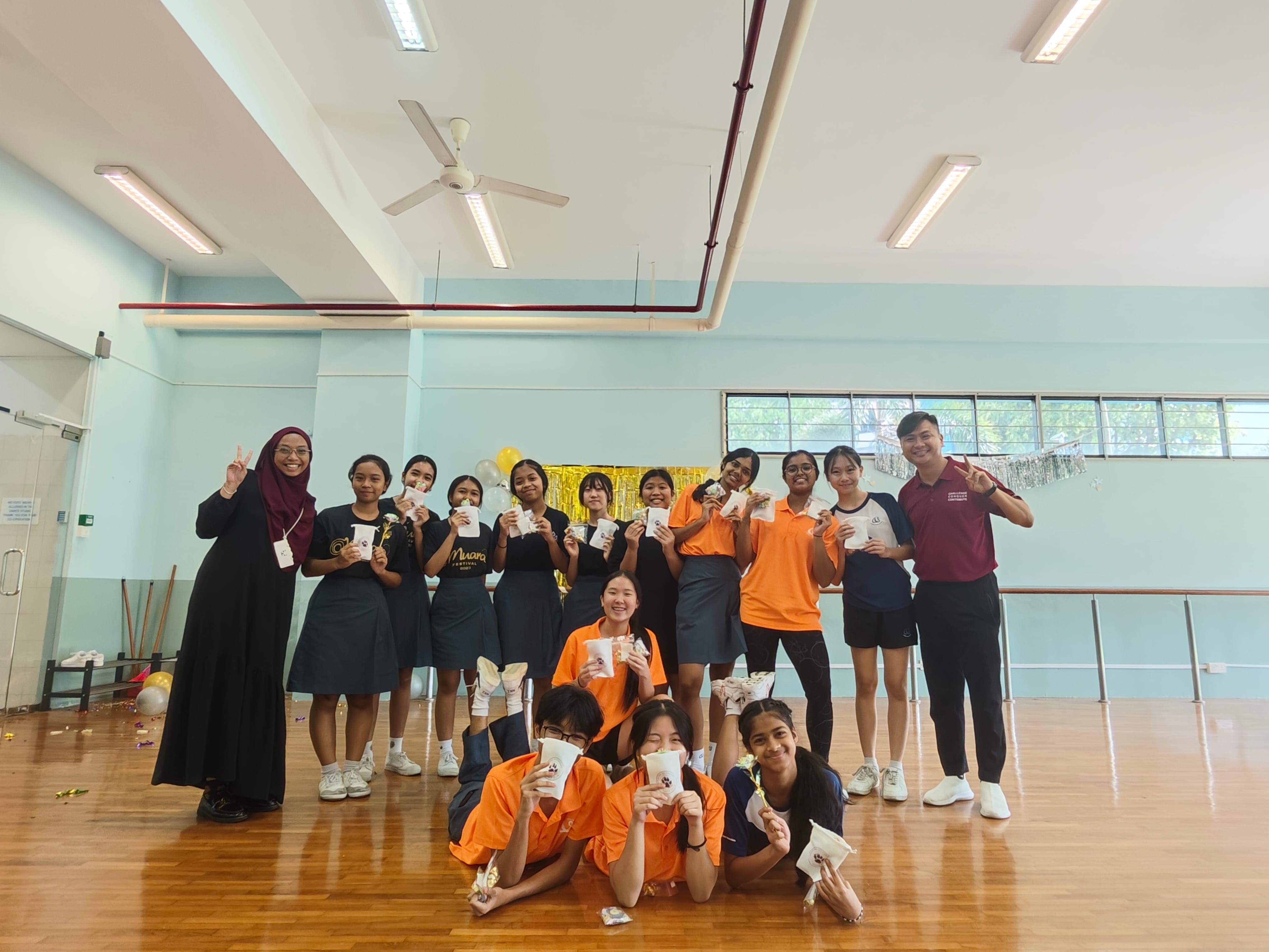 A group of students and two adults pose together in a school hall, holding cups and making peace signs.