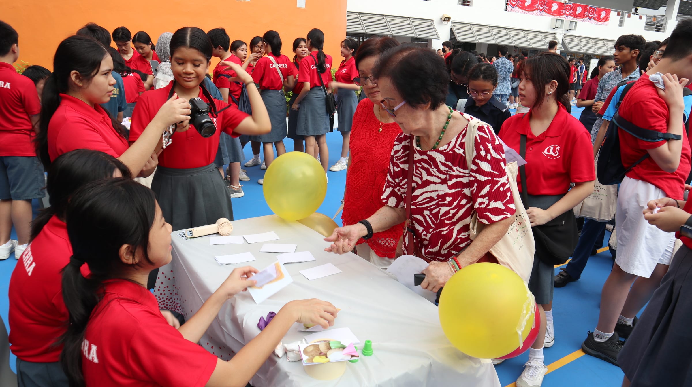 Students in red uniforms interact with elderly women at a table with yellow balloons during a school event.