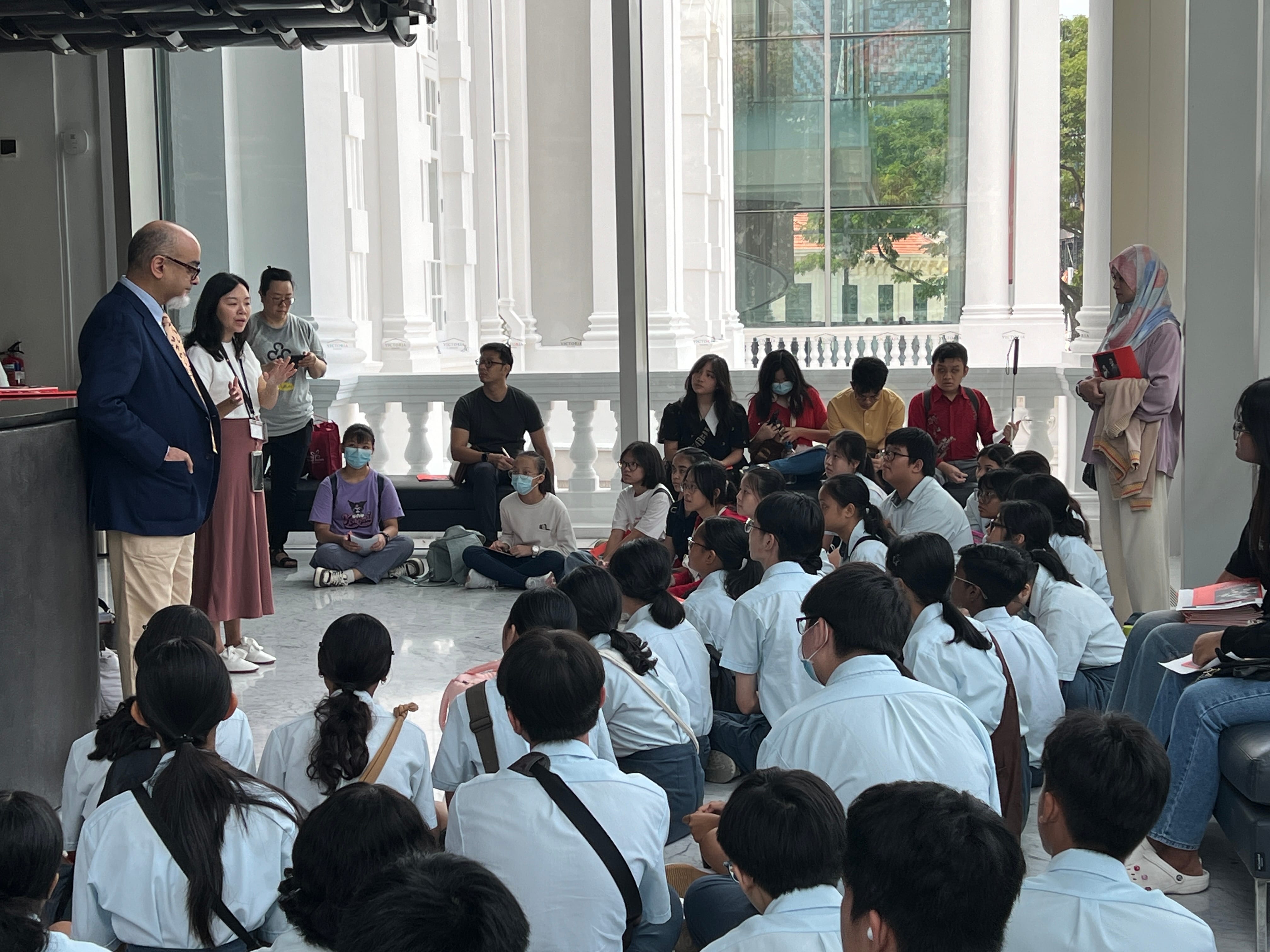 A group of students in white uniforms sit on the floor listening to two adults speaking in a bright building interior.