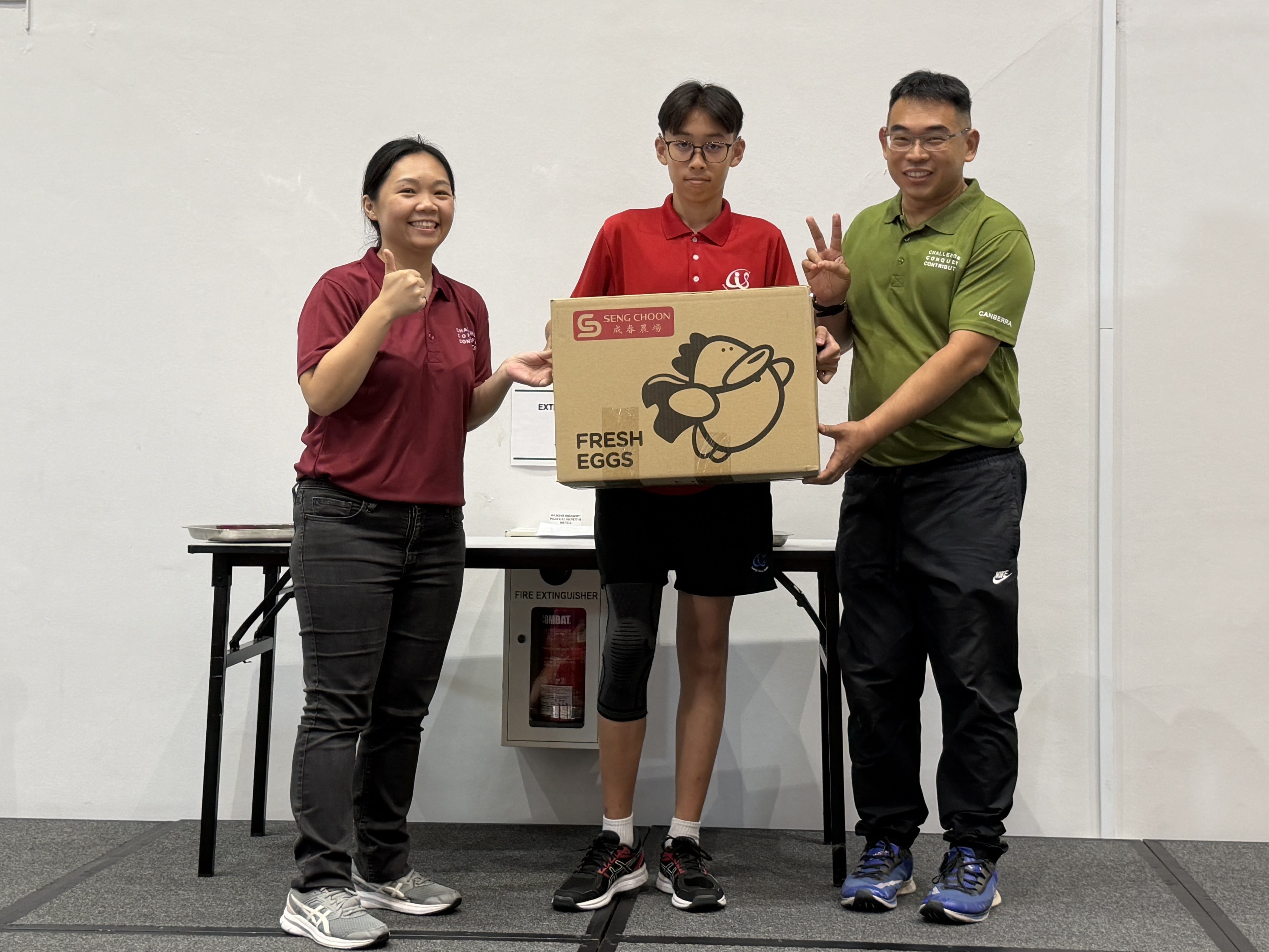 Two adults in maroon and green polo shirts flank a young male student in a red school polo shirt, all three holding a Seng Choon Fresh Eggs cardboard box.