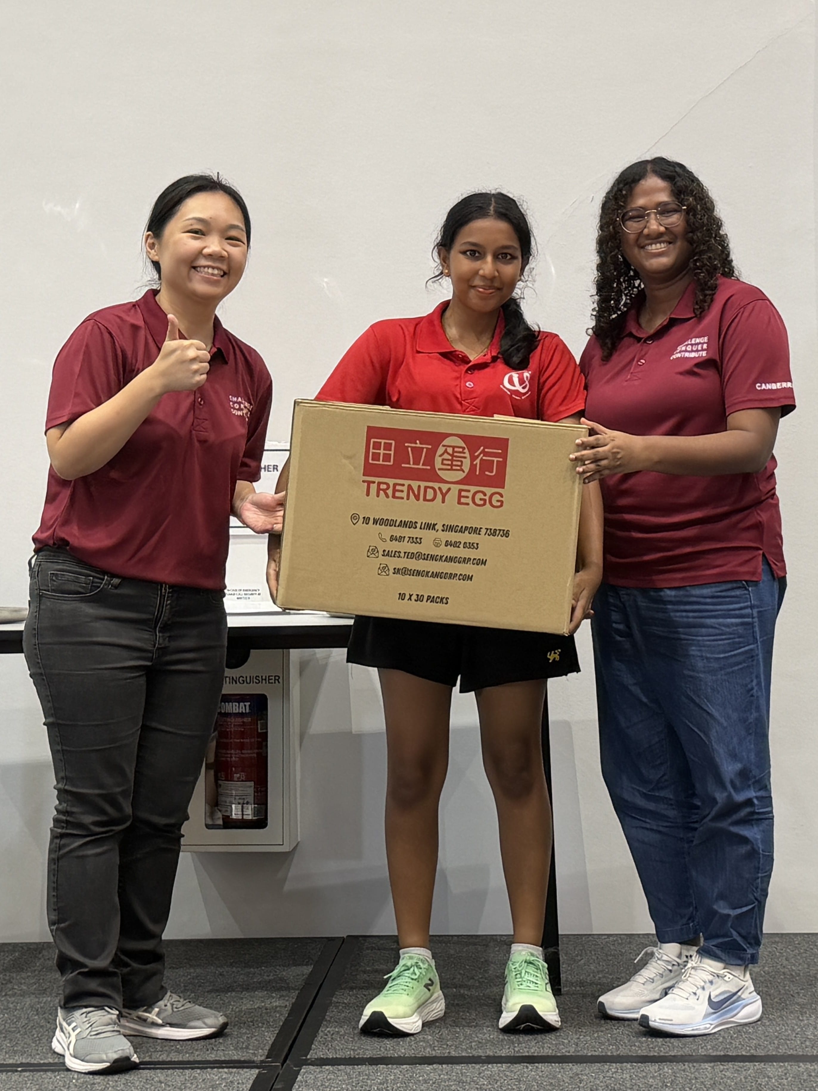 Three people in maroon polo shirts pose together holding a large cardboard box from Trendy Egg (田立蛋行), a Singapore egg supplier. The person on the left gives a thumbs up.