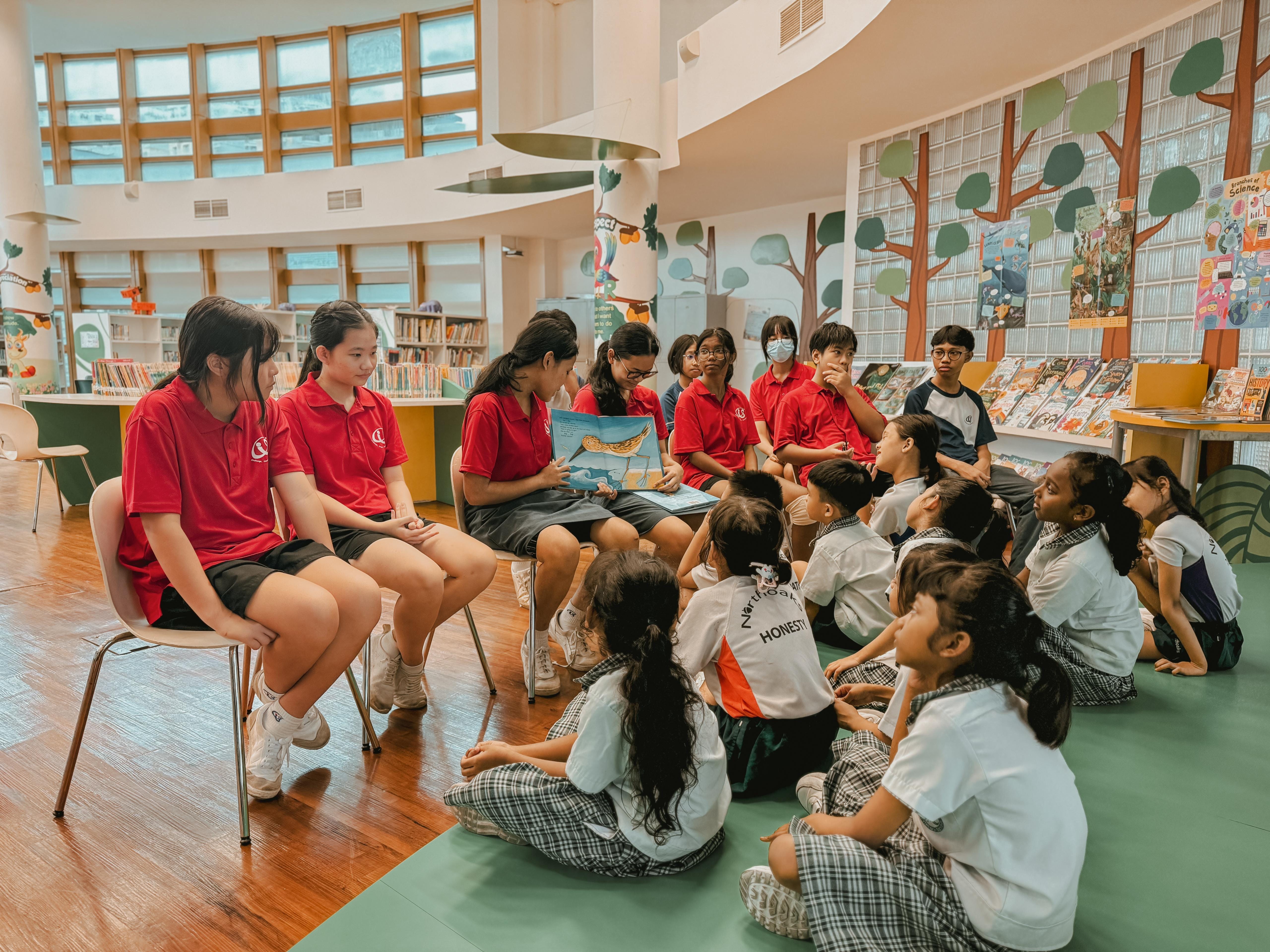 A group of secondary school students wearing red tees read a book to a group of primary school students sitting on the floor in front of them