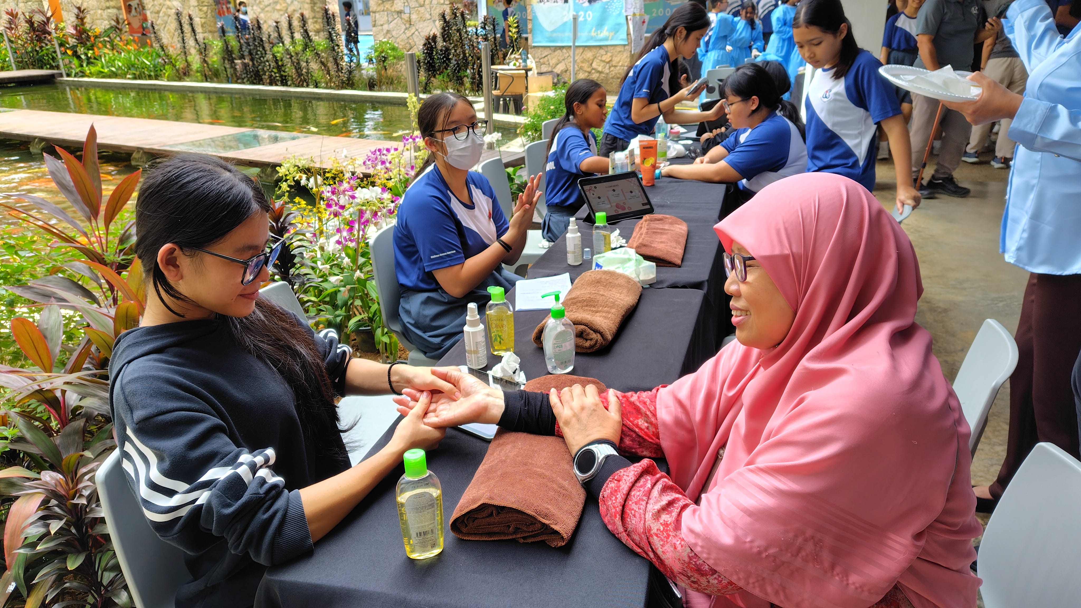 A woman in a pink hijab gives a hand massage to a young woman at an outdoor wellness booth with students nearby.