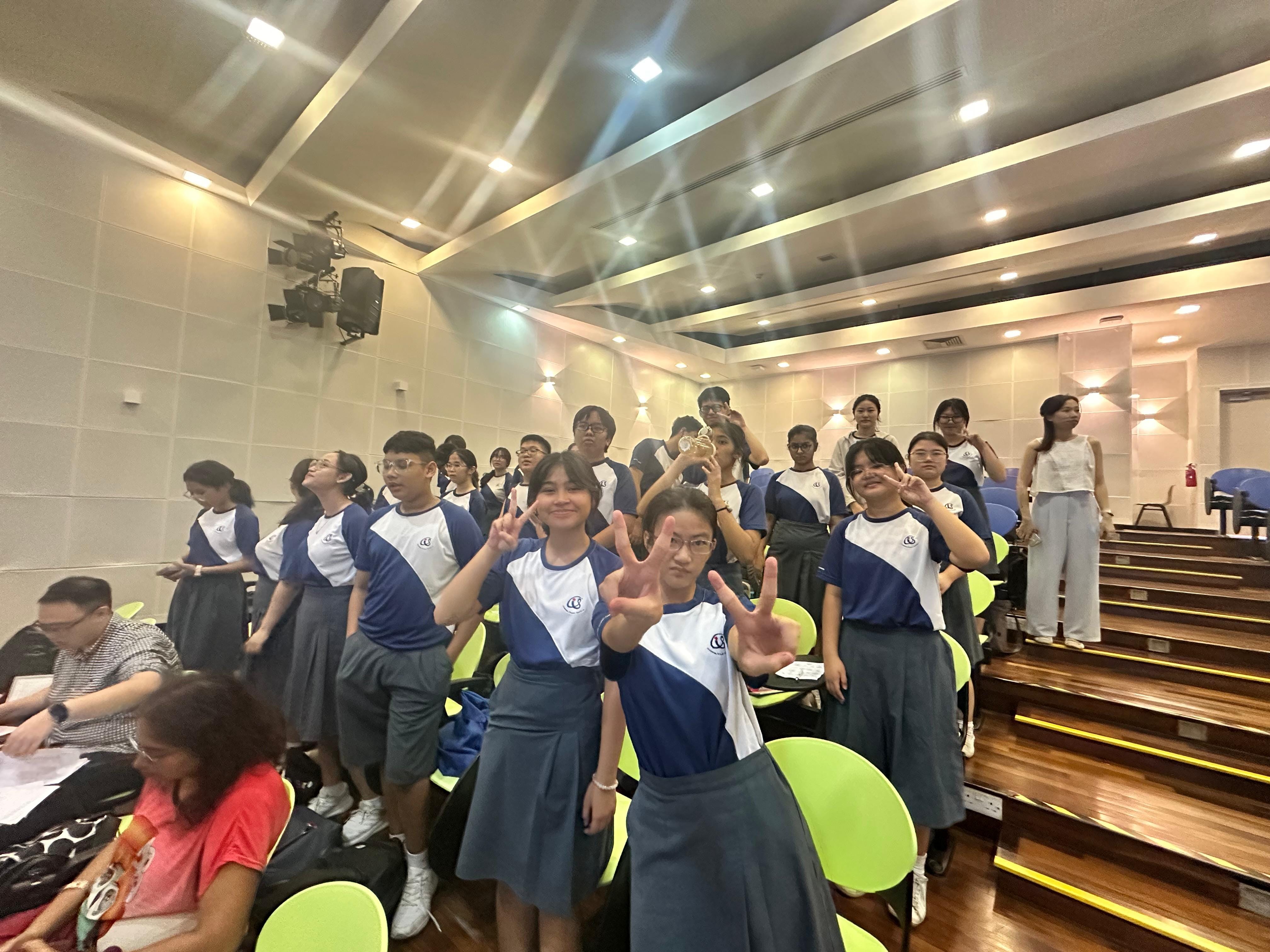 Students in blue and white uniforms pose with peace signs in a tiered auditorium with green chairs.