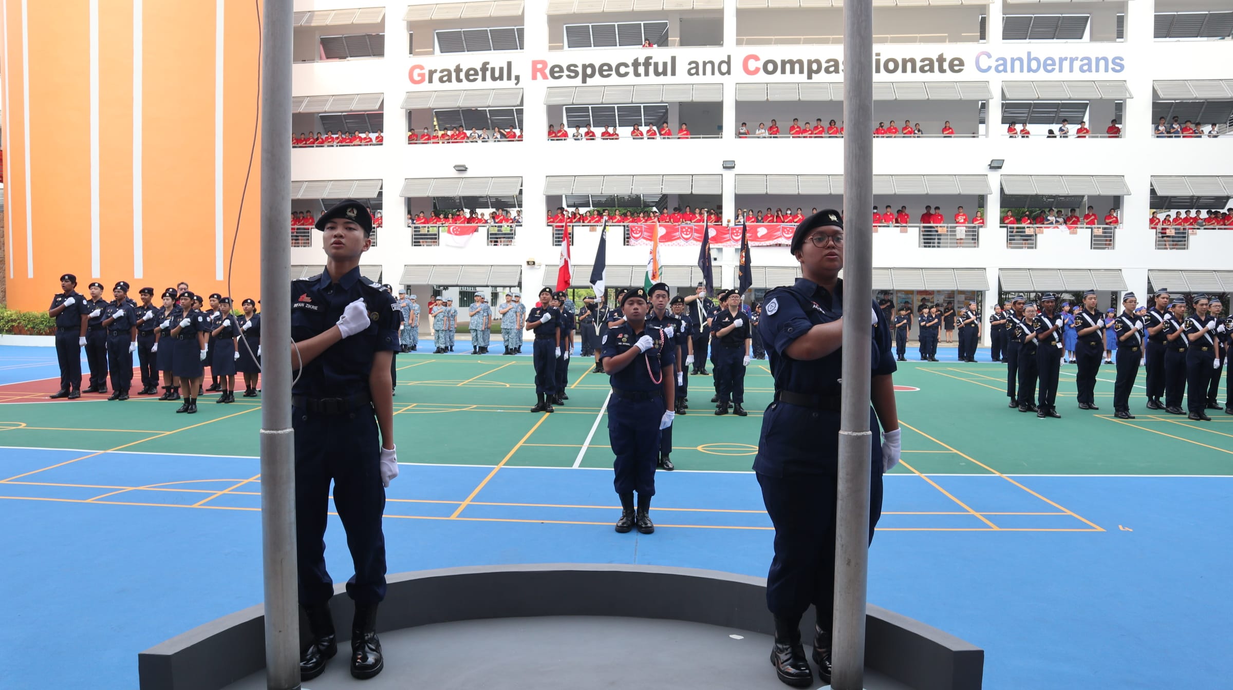 Students in navy uniforms stand at attention during a school parade at Canberra Secondary, with spectators on balconies above.