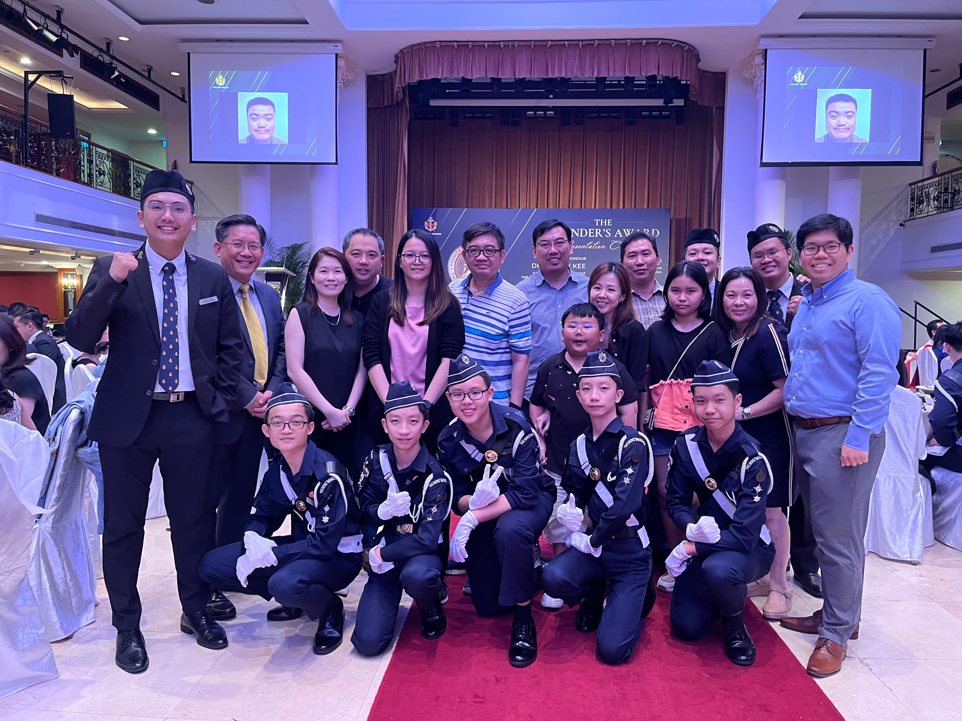 Group of adults and youth in dark uniforms posing together at a formal awards ceremony in a decorated hall.