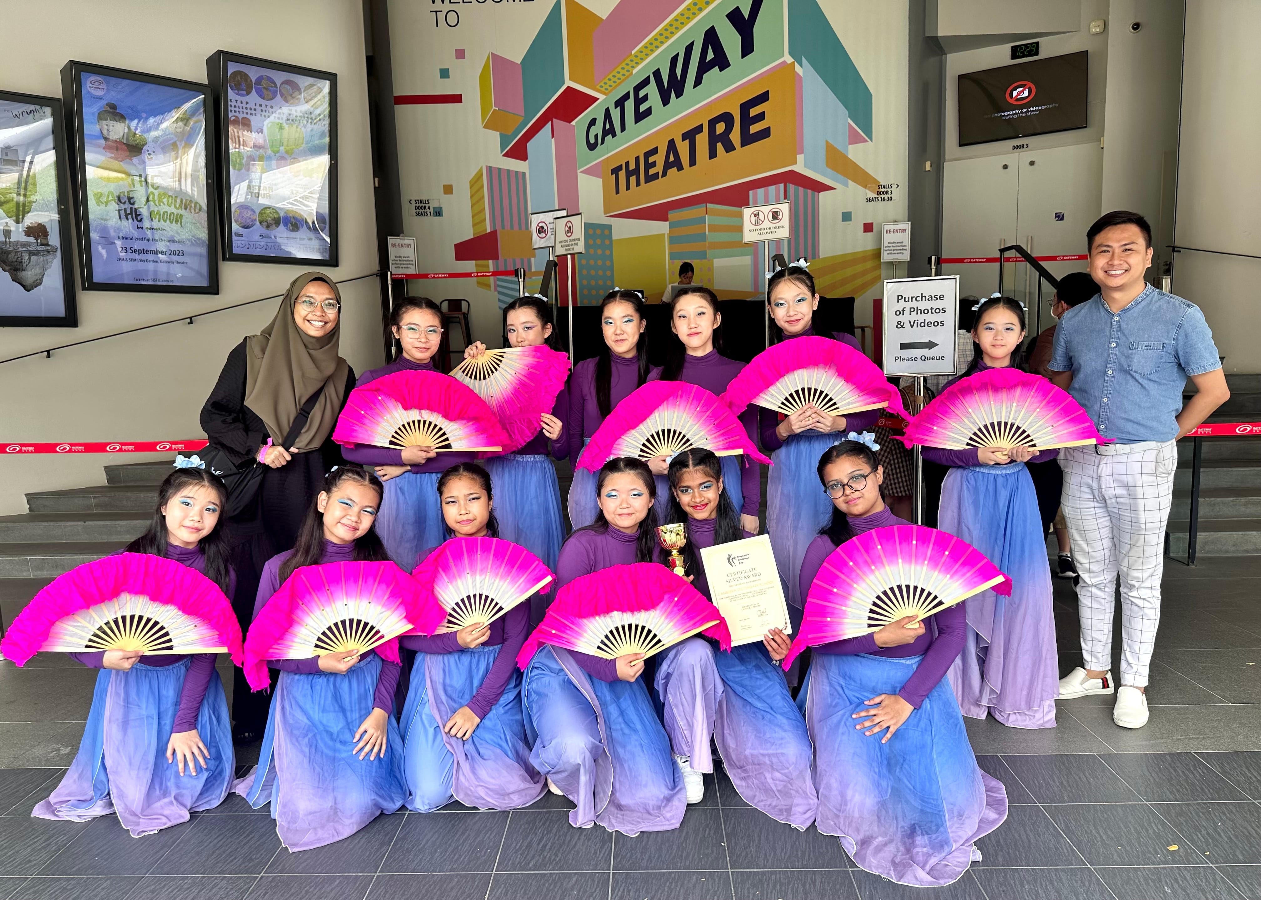 Young dancers in purple costumes holding pink fans pose with two adults outside Gateway Theatre, one holding a trophy and certificate.