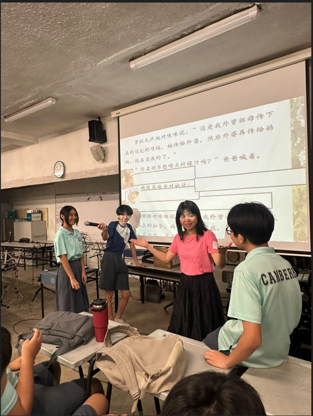 A teacher in a pink t-shirt facilitates a student activity at the front of a classroom. Two students — one holding a microphone — stand beside her in front of a projector screen displaying a Chinese text passage. A student in a Canberra Secondary School uniform watches from the foreground.