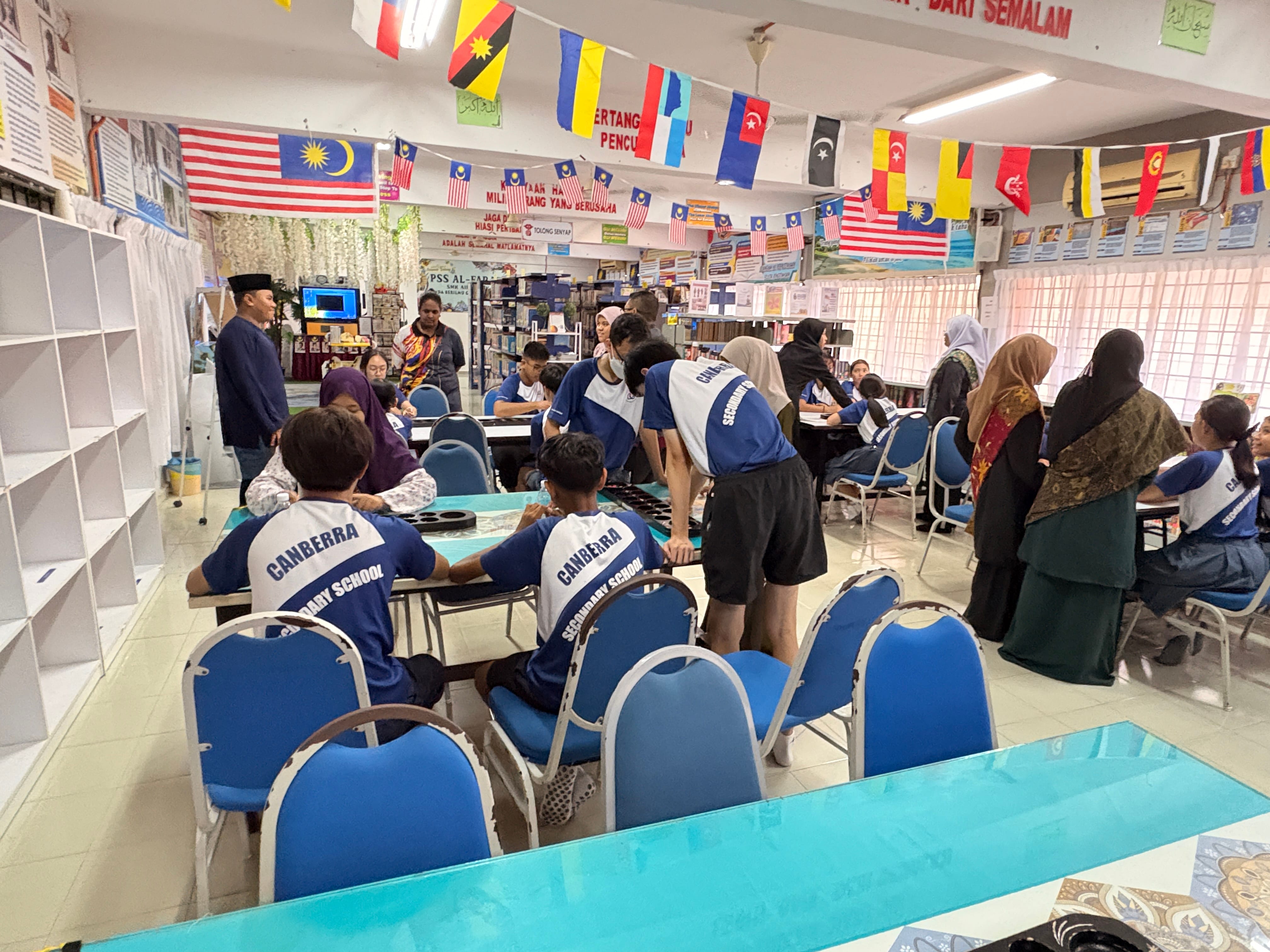 Students in Canberra Secondary School uniforms sit and stand at tables in a room decorated with Malaysian flags.