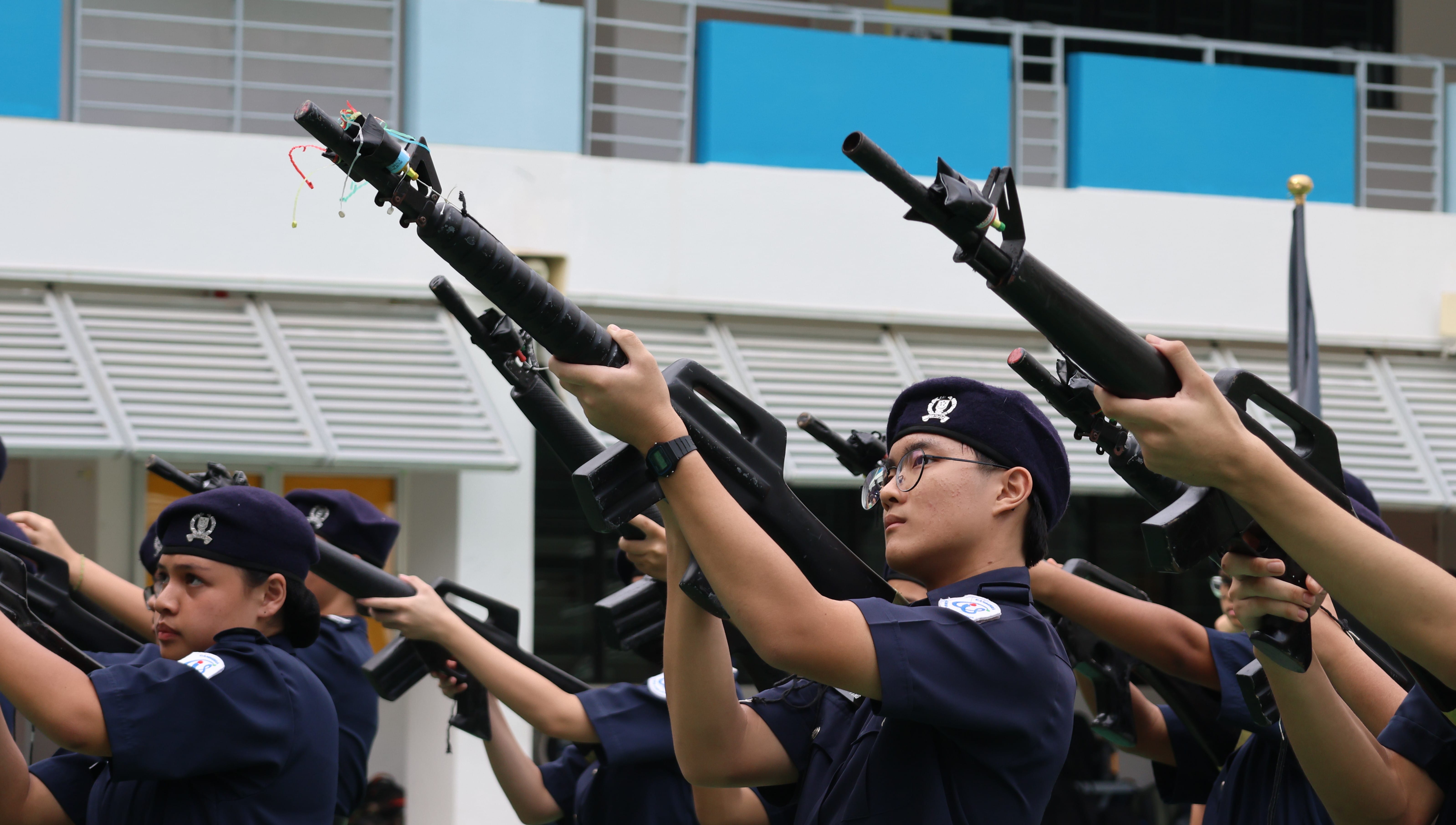 Young cadets in navy uniforms and berets raise rifles overhead during an outdoor drill formation.