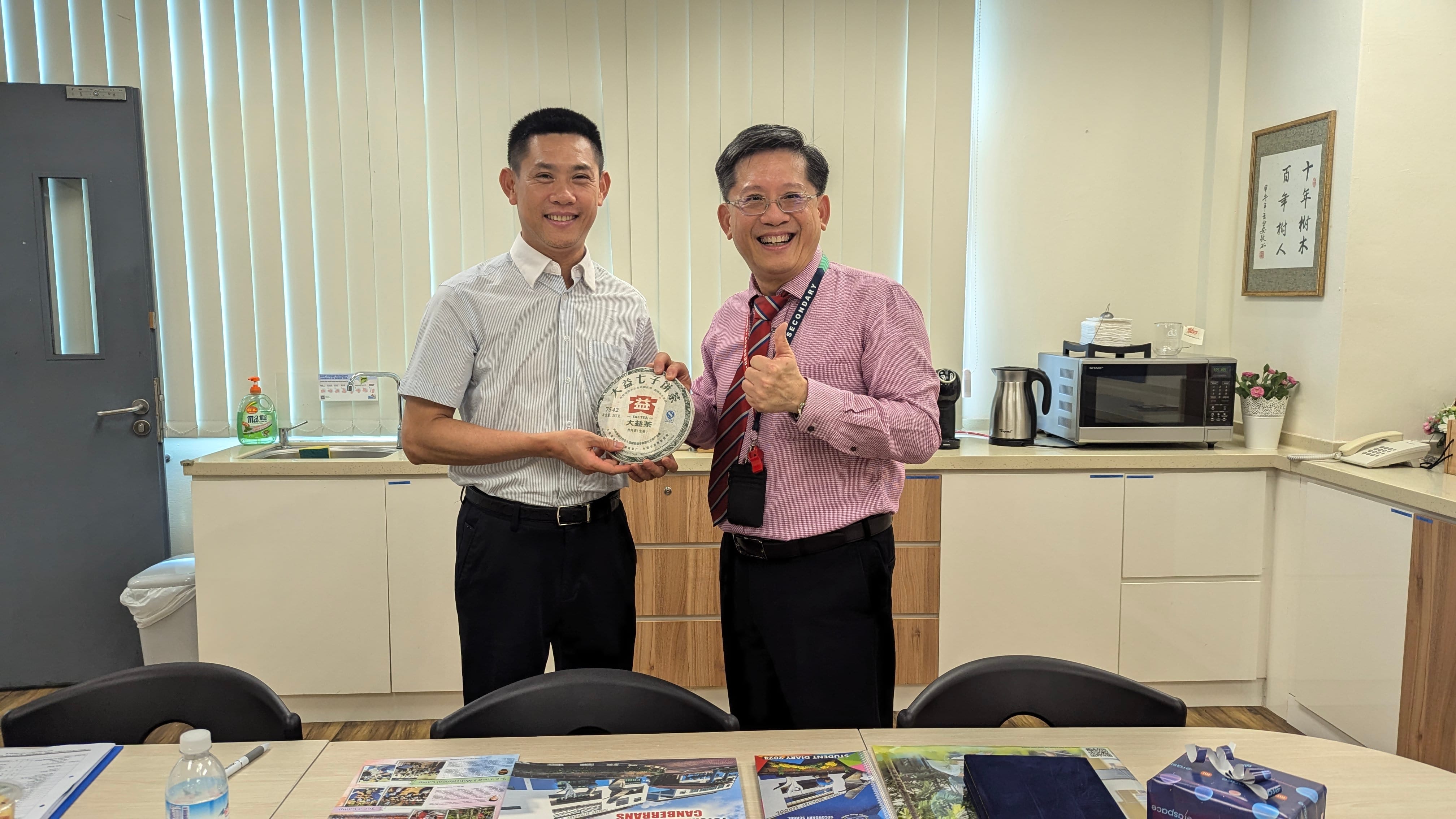 Two smiling men stand together in an office, holding a round Chinese tea cake, with one giving a thumbs up.