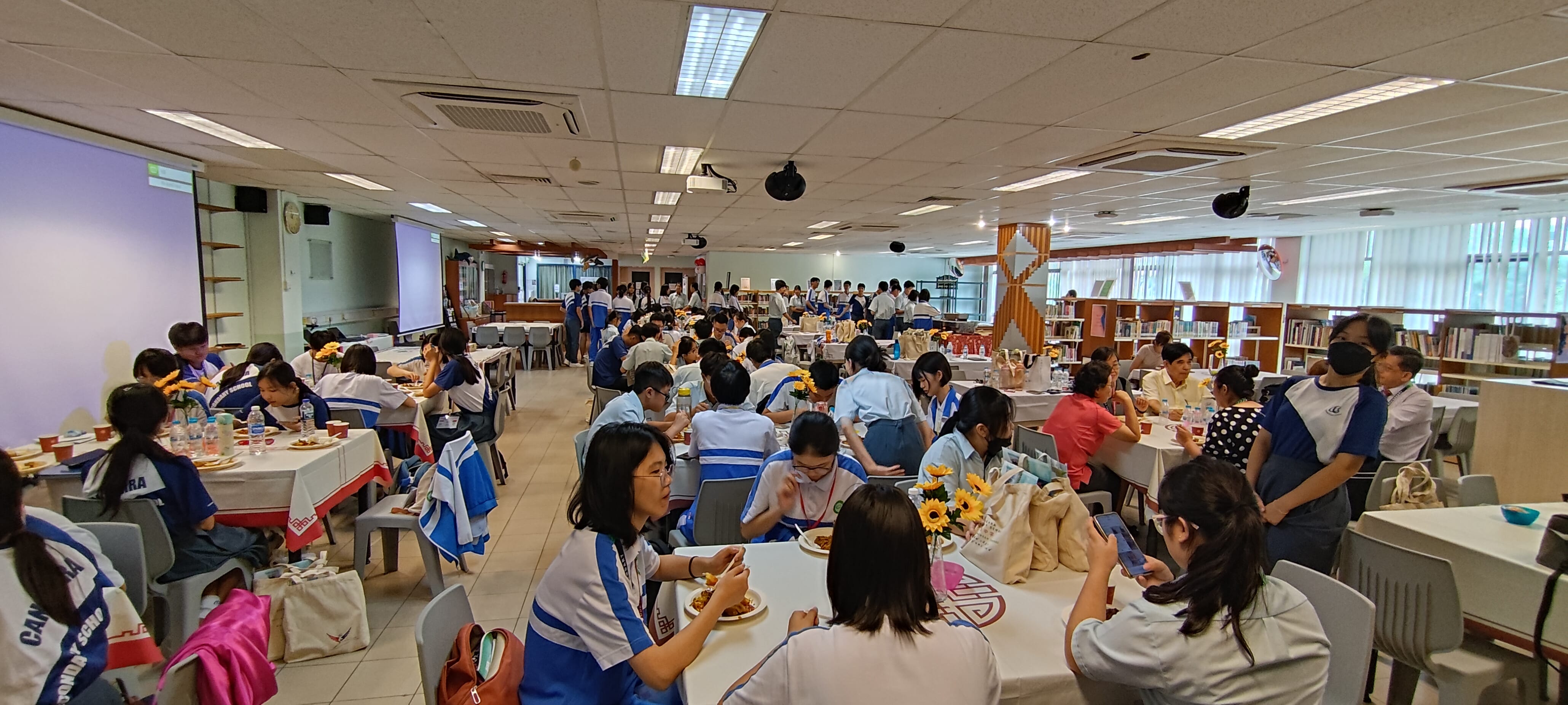 Students in blue and white uniforms sit at round tables with sunflowers in a large school hall during an event.