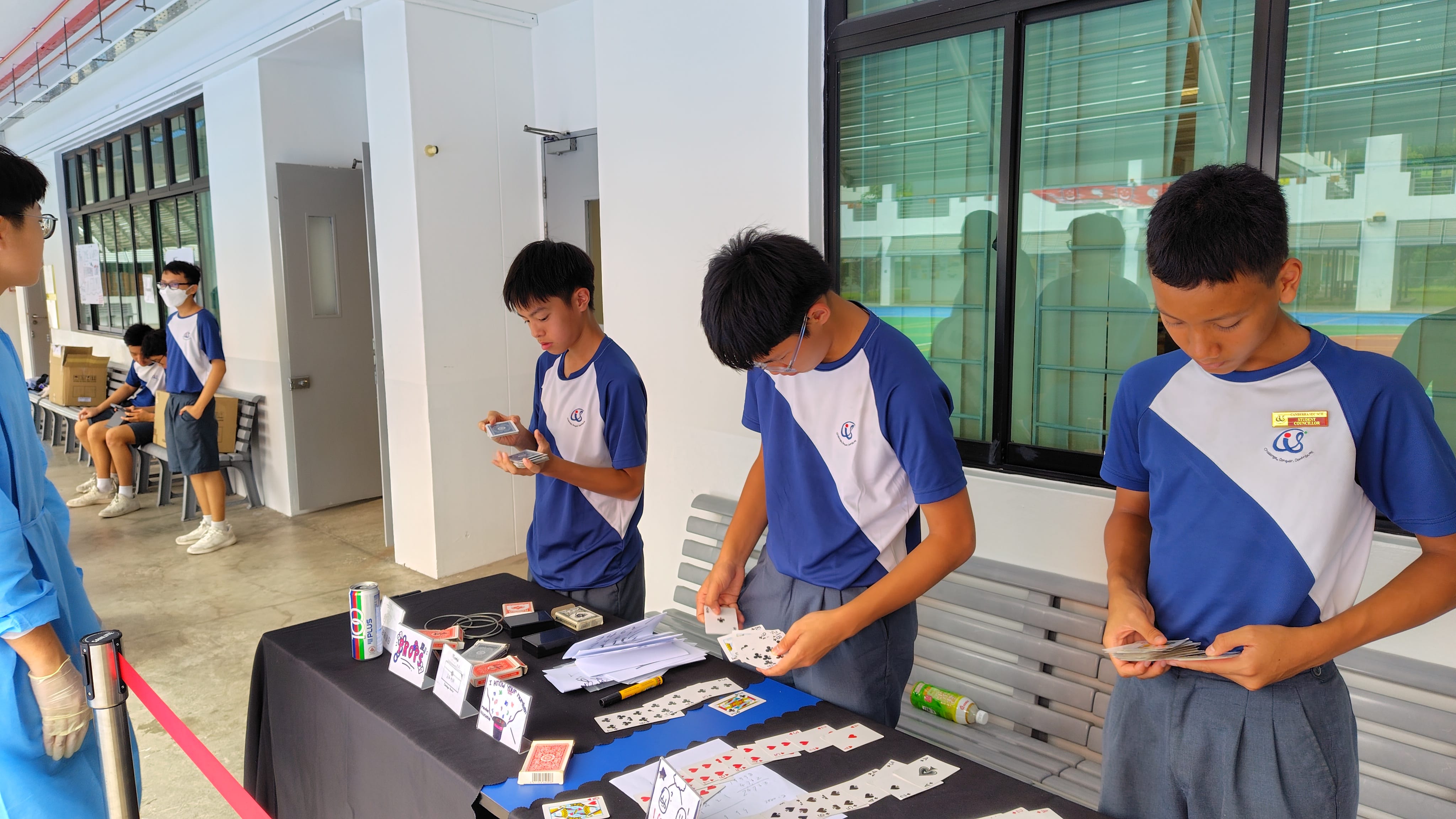 Three boys in blue school uniforms handle playing cards at a table covered with cards and magic props.