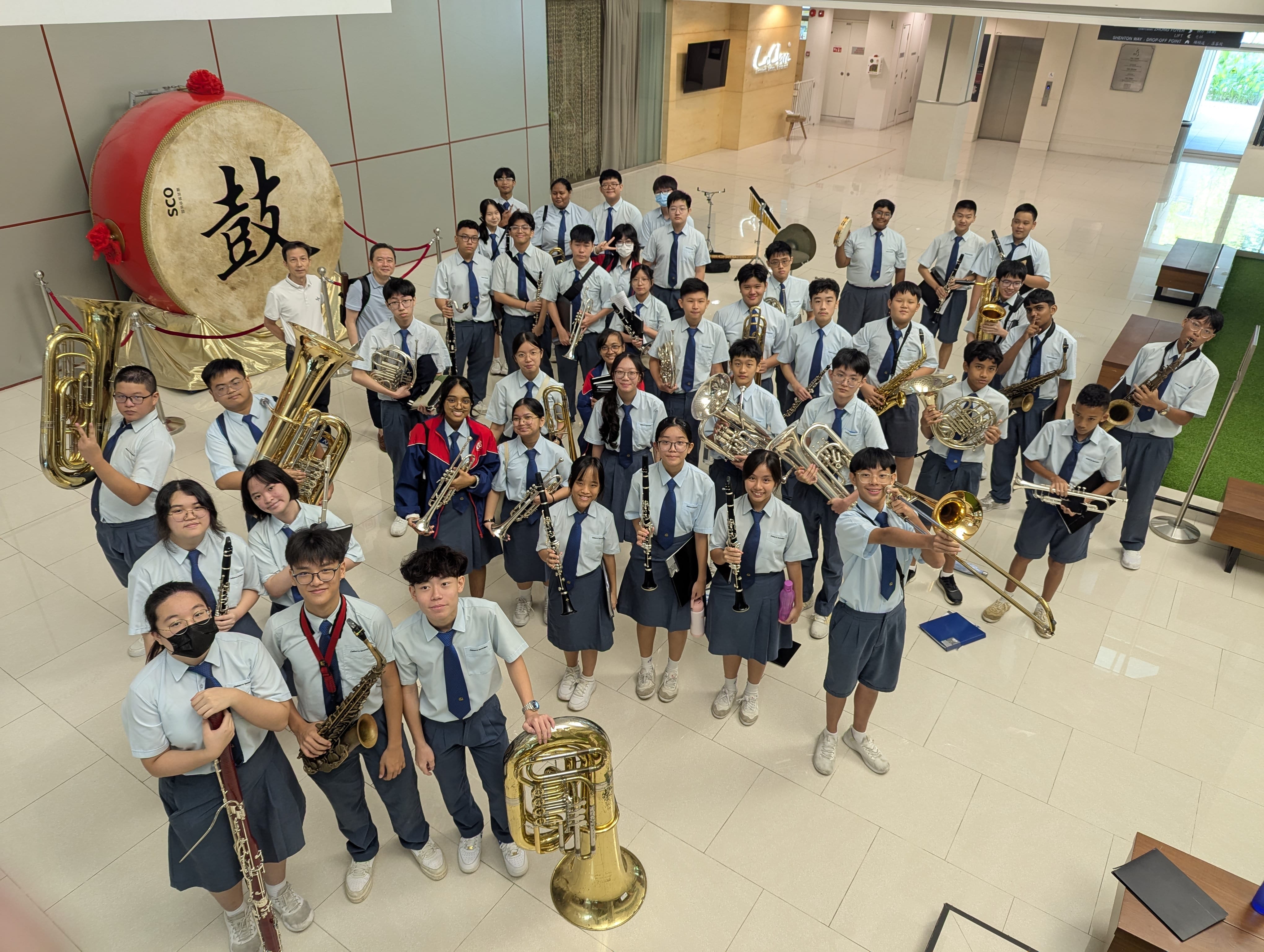 A large school band in uniforms holds brass and woodwind instruments indoors near a large red Chinese drum.