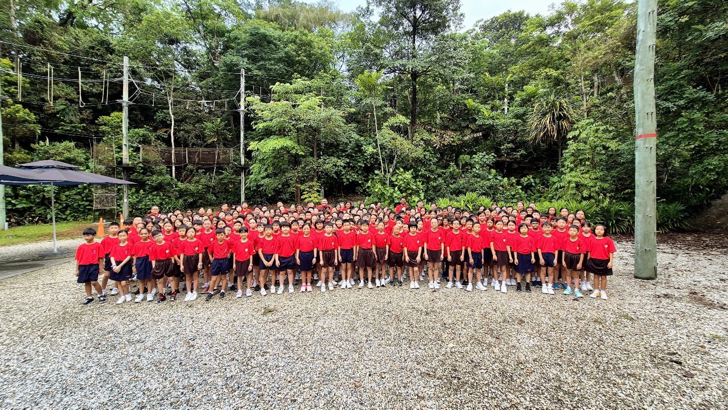 Large group of children in red shirts and dark shorts standing on gravel outdoors with dense green trees behind them.