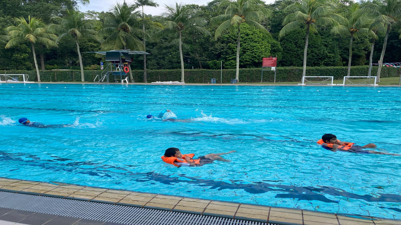Children swimming in an outdoor pool, two wearing orange life vests, with palm trees and a lifeguard stand in the background.