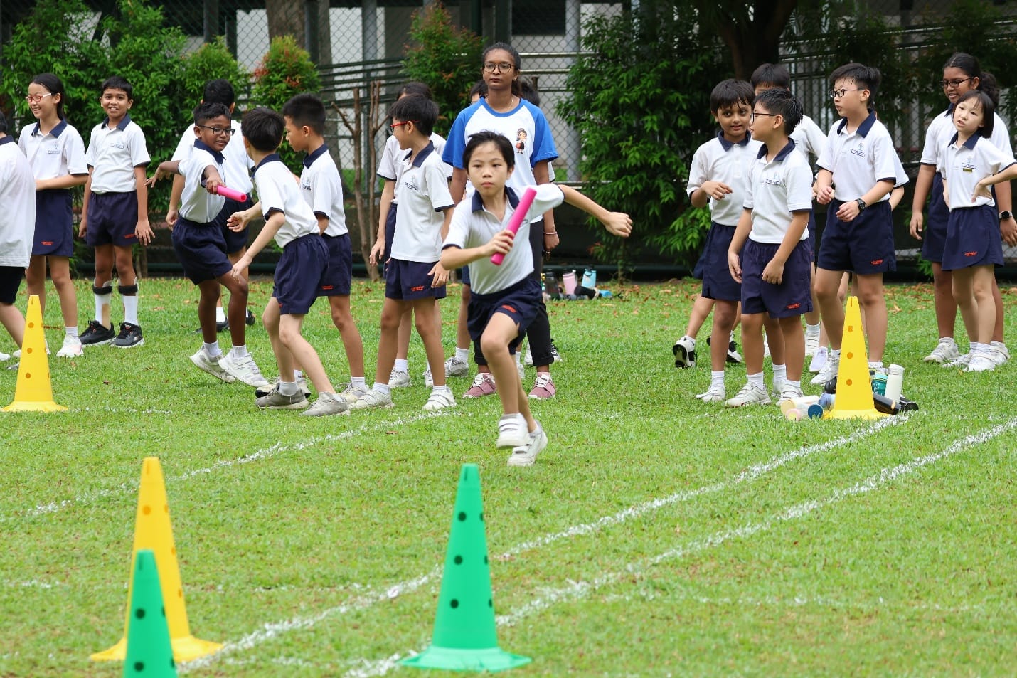 Student runs holding a pink baton during a relay race on grass, with classmates and cones nearby.