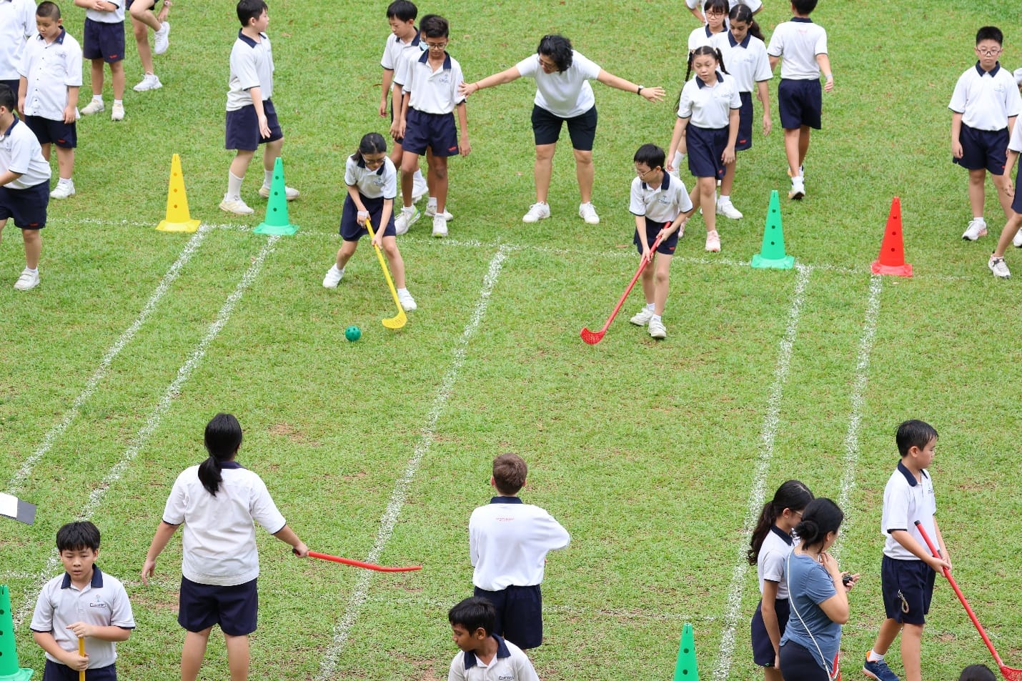 Students play field hockey on a grass field with colorful cones and a teacher supervising.