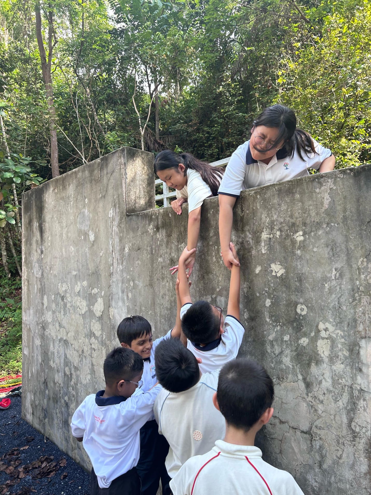 Students help each other climb over a tall concrete wall outdoors, surrounded by trees.