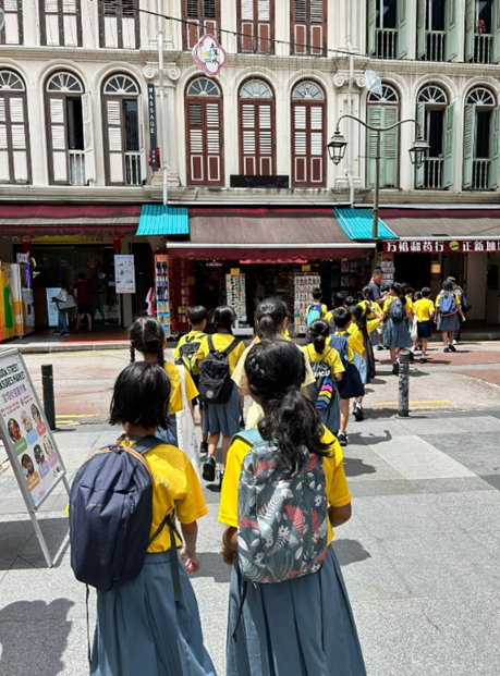 Children in yellow shirts and gray skirts with backpacks walk toward a row of shophouses with Chinese signage.