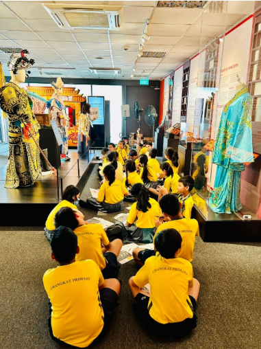 Students in yellow uniforms sit on a museum floor facing traditional costume displays and mannequins.