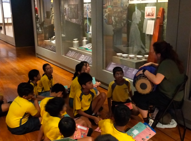 Children in yellow uniforms sit on a museum floor listening to a guide holding a drum near display cases.