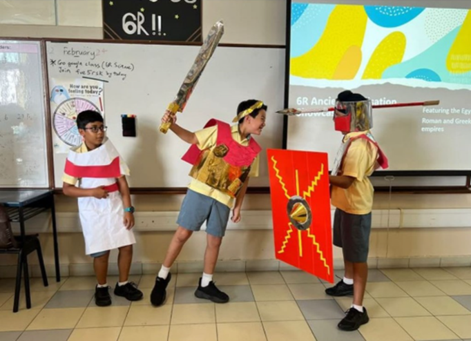 Three students in handmade ancient Roman costumes with cardboard sword and shield present in a classroom.