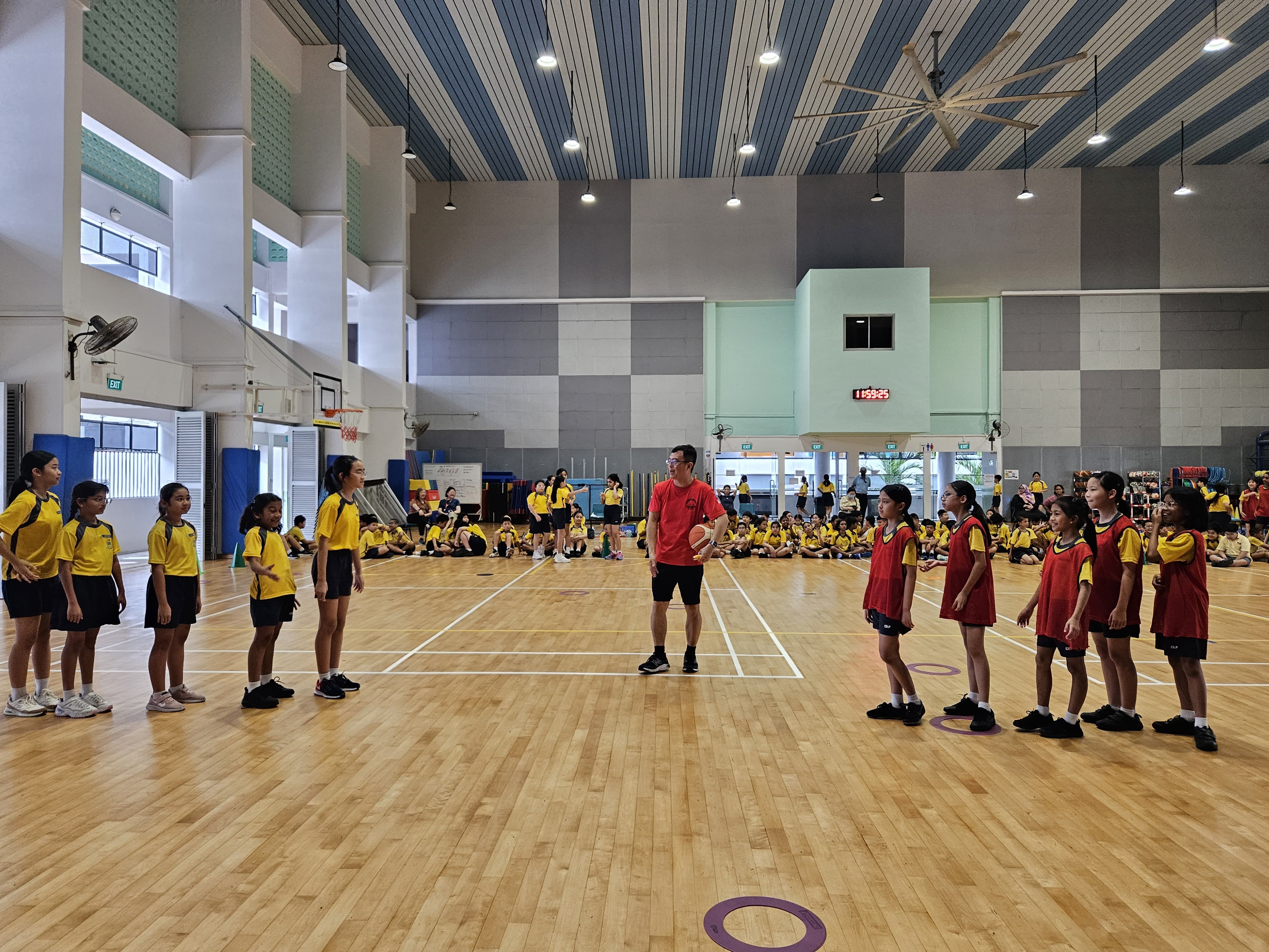 A coach holding a basketball stands between two lines of children in yellow and red jerseys in a school gymnasium.