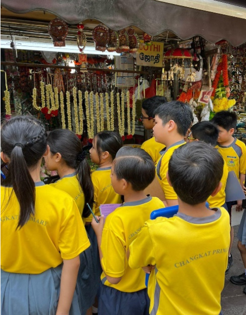 Children in yellow Changkat Primary shirts look at a shop with hanging flower garlands and decorations.