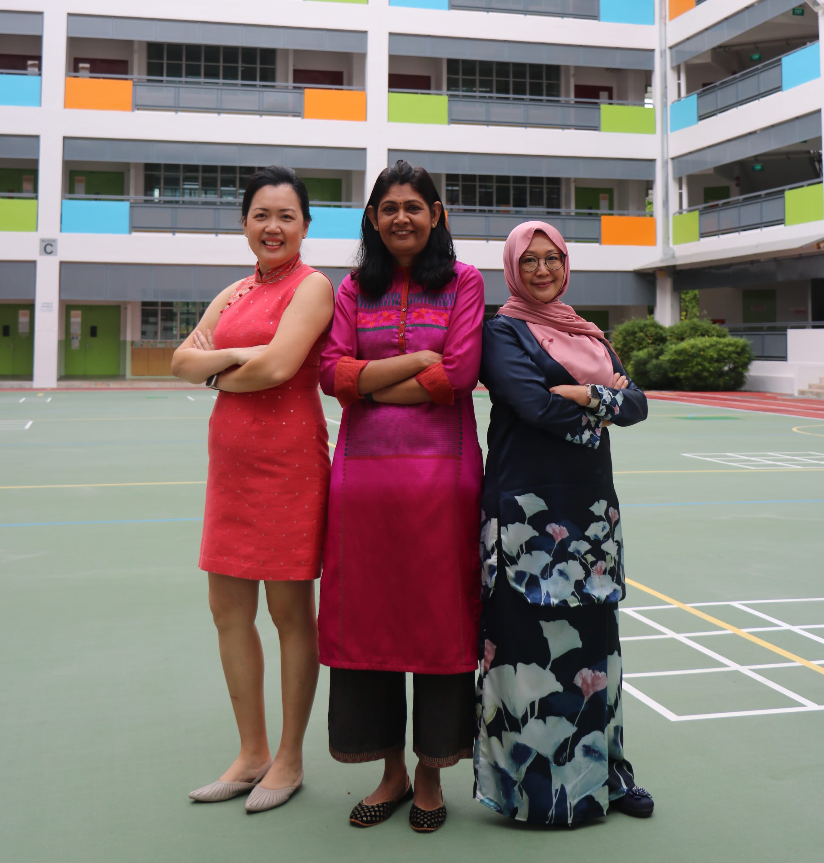 Three women in traditional attire stand arms crossed on a school courtyard, smiling, with a colorful multi-storey building behind them.