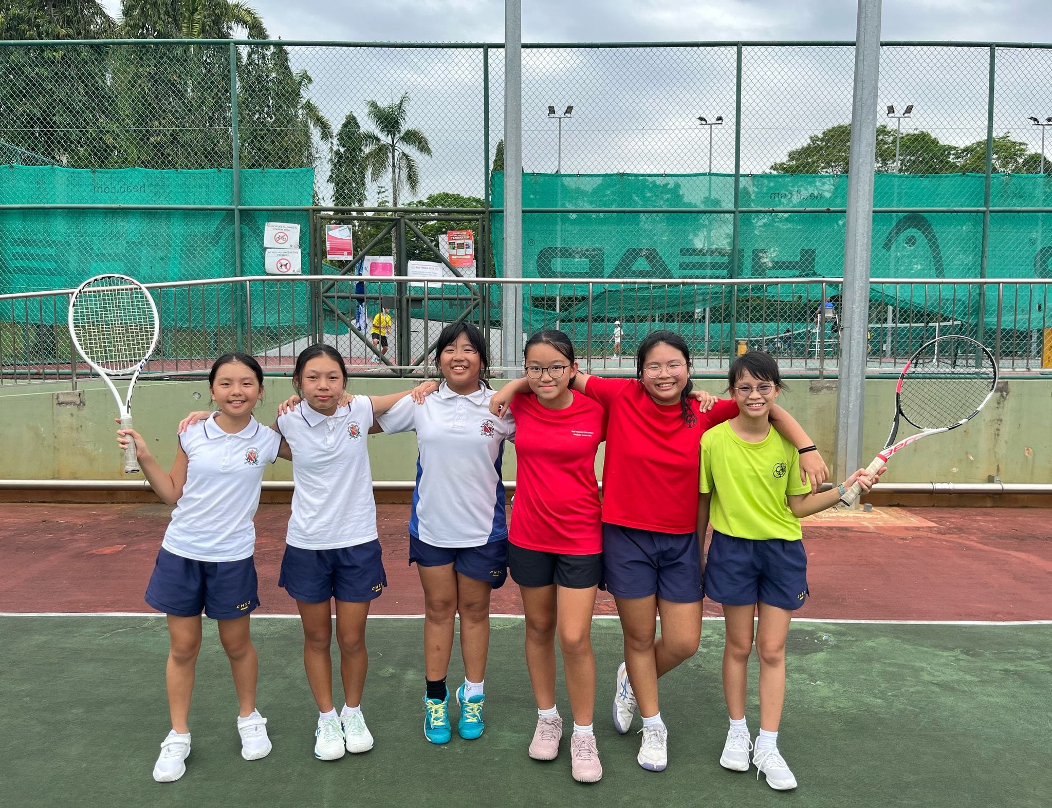 Six young girls in sports uniforms pose together smiling on a tennis court, two holding rackets.
