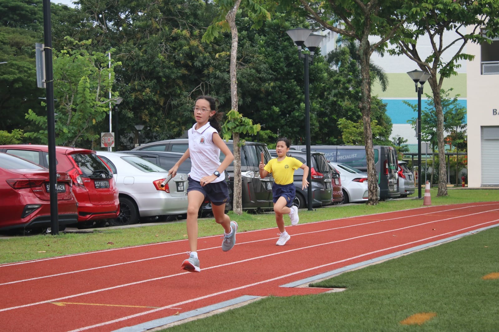 Two young girls run a relay race on a red track, each holding a baton, with parked cars and trees behind them.