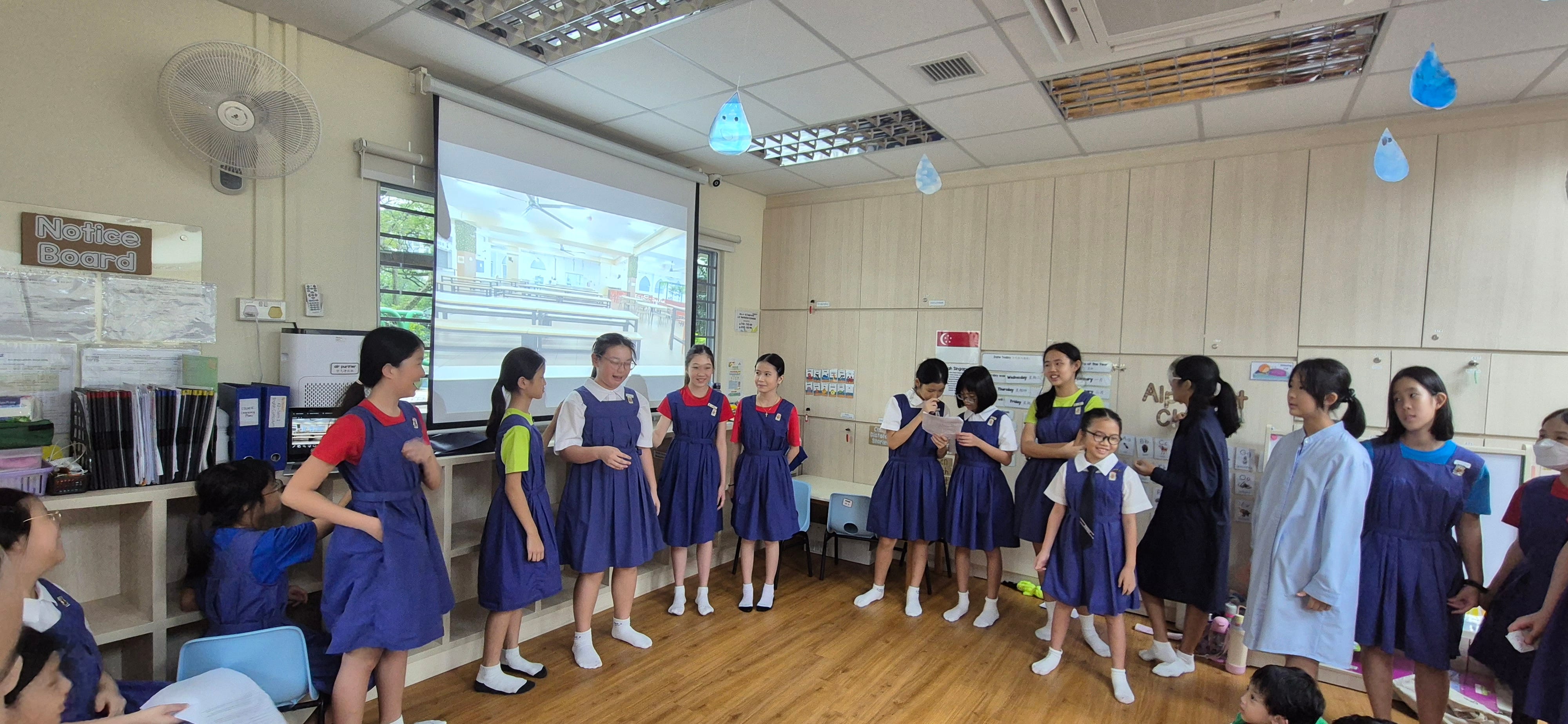Girls in blue school uniforms stand in a semicircle in a decorated classroom with a projector screen behind them.