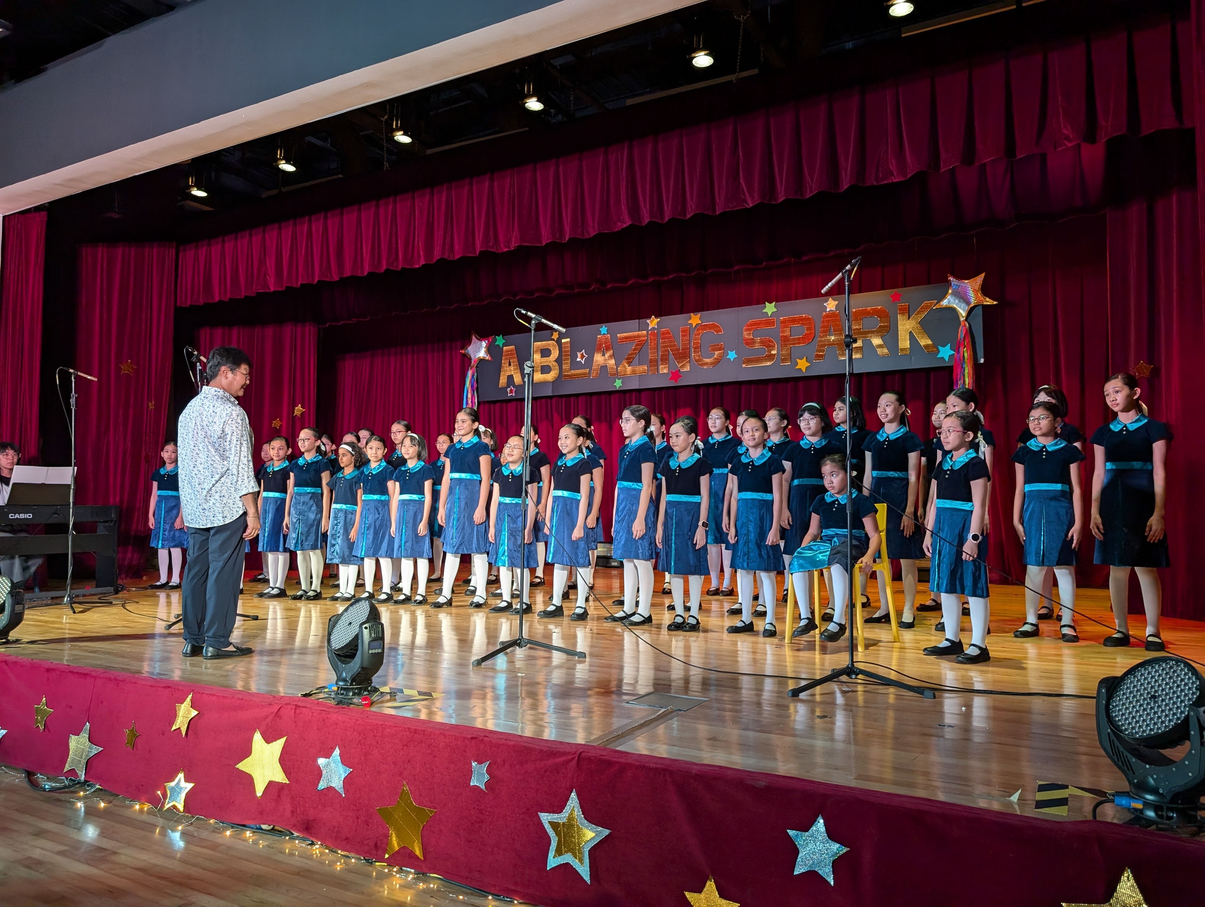 A children's choir in blue uniforms performs on a decorated stage under a sign reading "A Blazing Spark."
