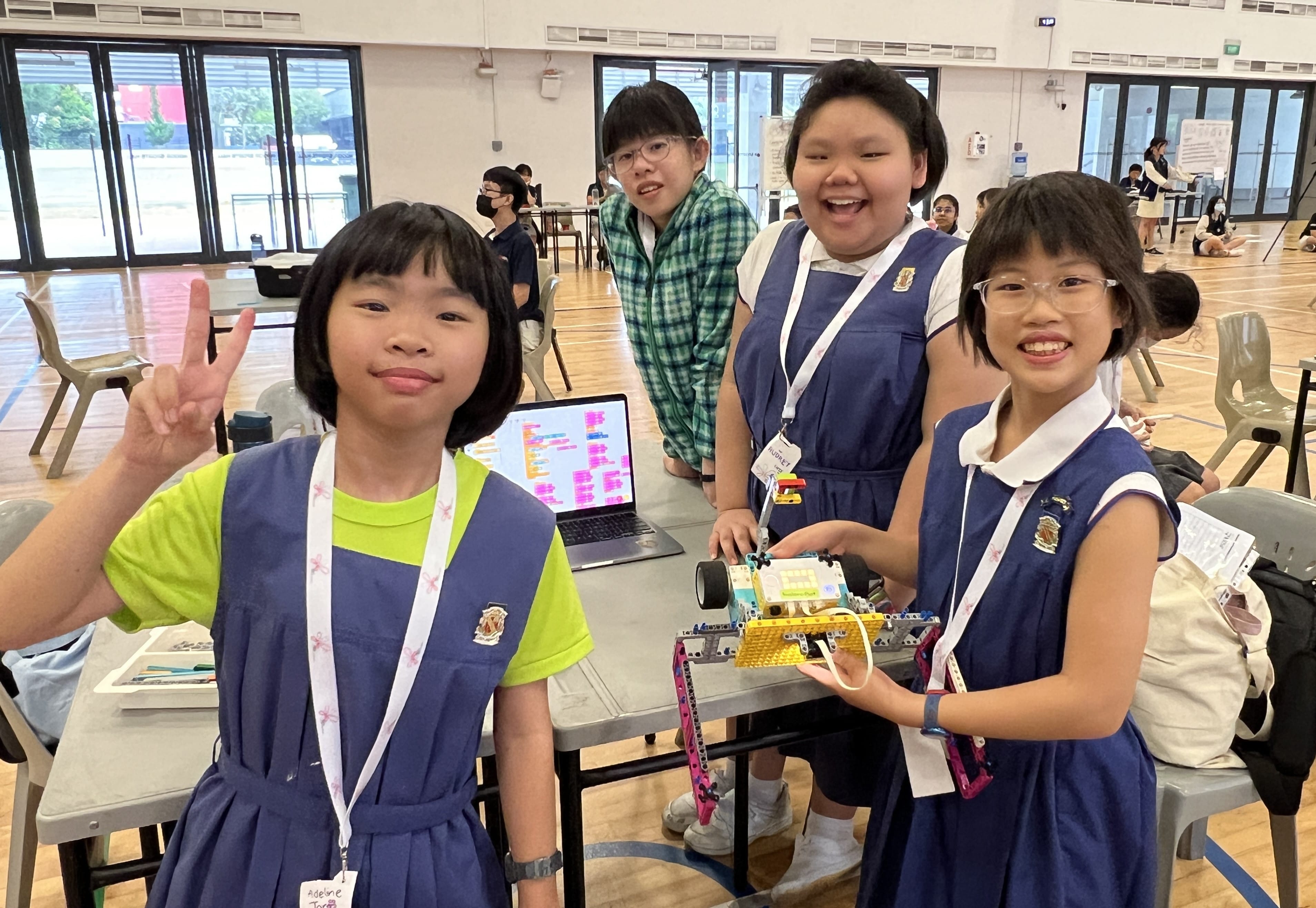 Four children in school uniforms smile at a table with a LEGO robot and a laptop showing a coding program.