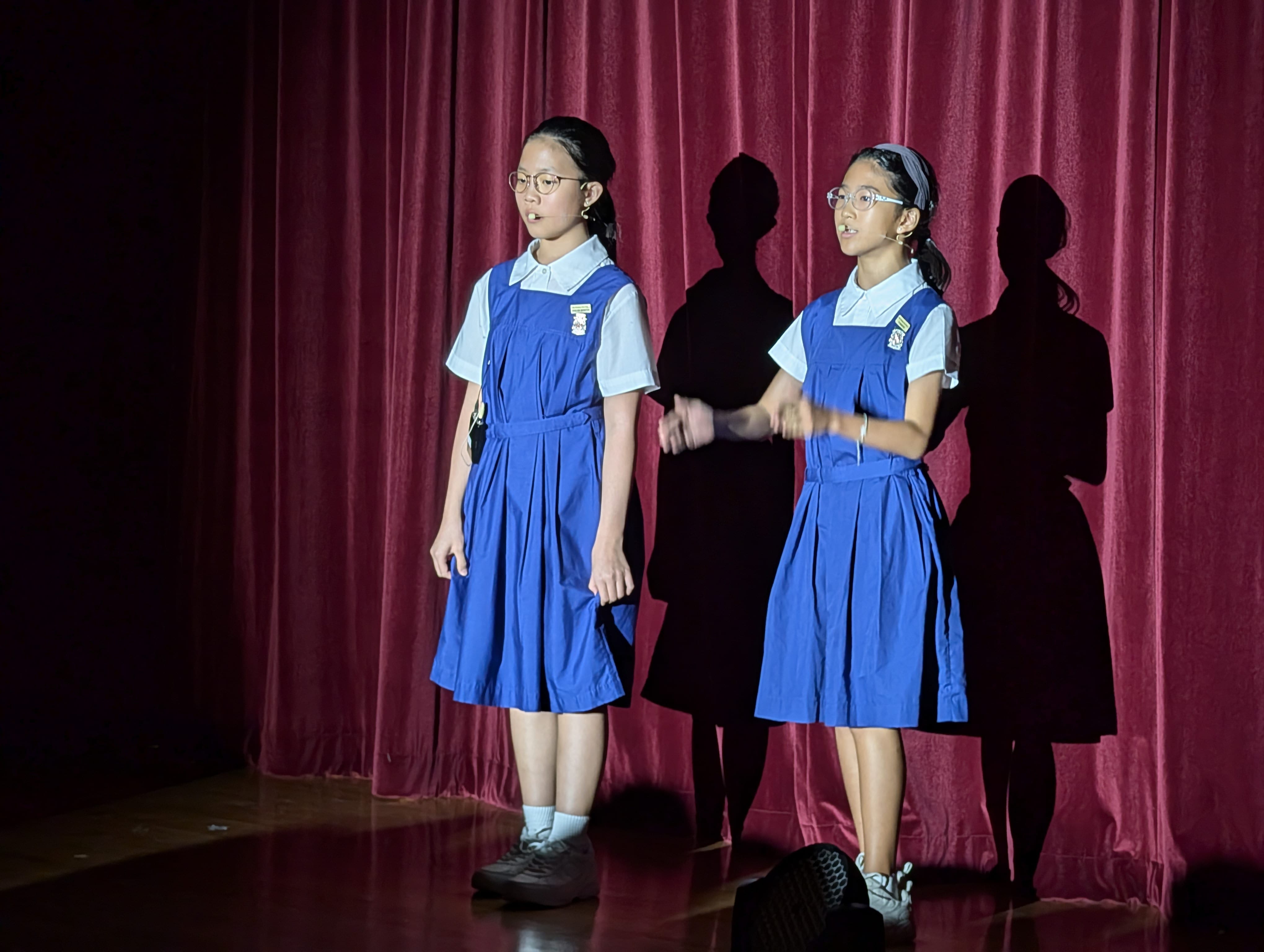 Two girls in blue school uniforms and glasses perform on a stage with a red curtain backdrop.