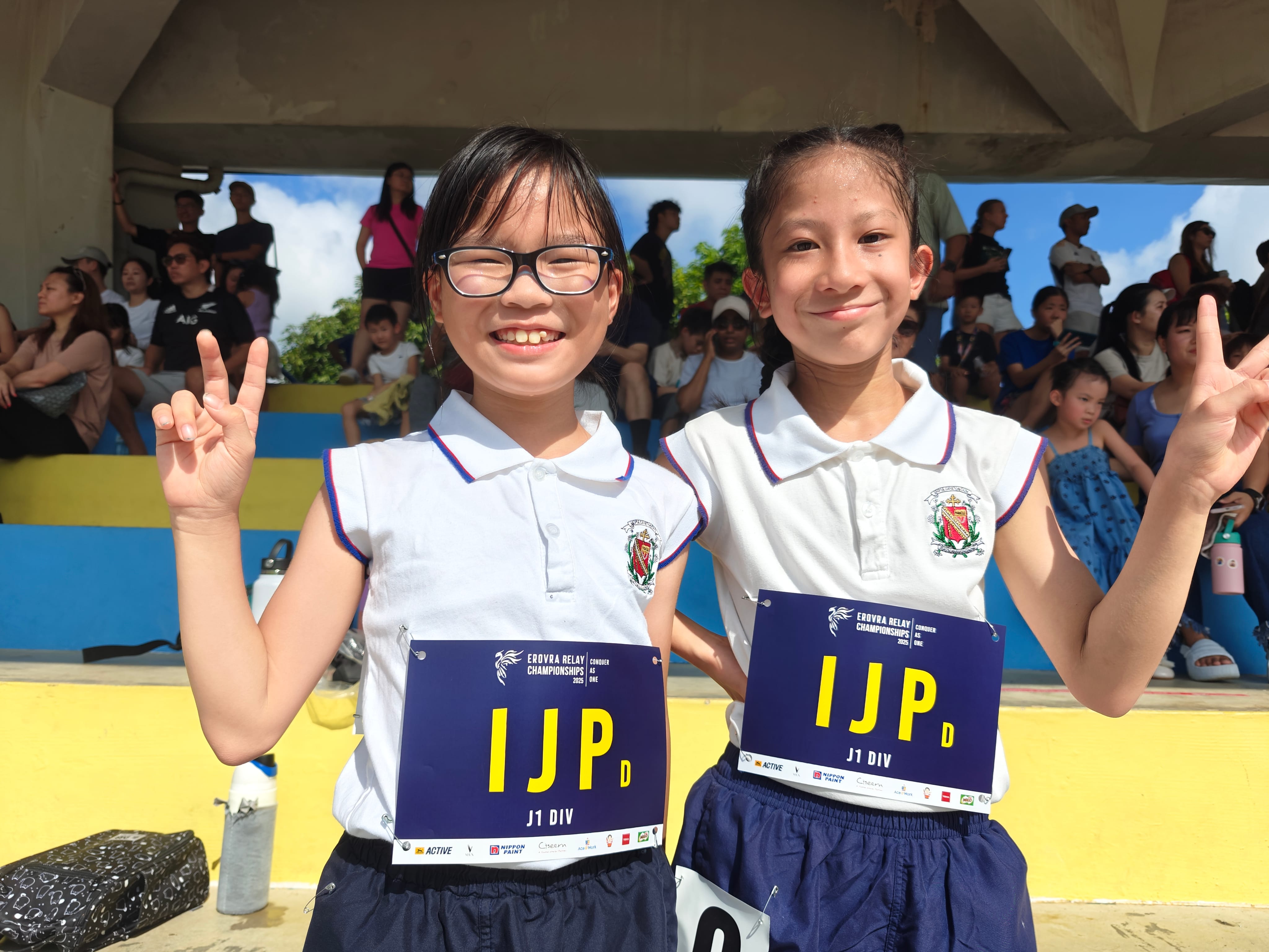 Two smiling girls in sports uniforms show peace signs, with race bibs, and spectators on bleachers behind.