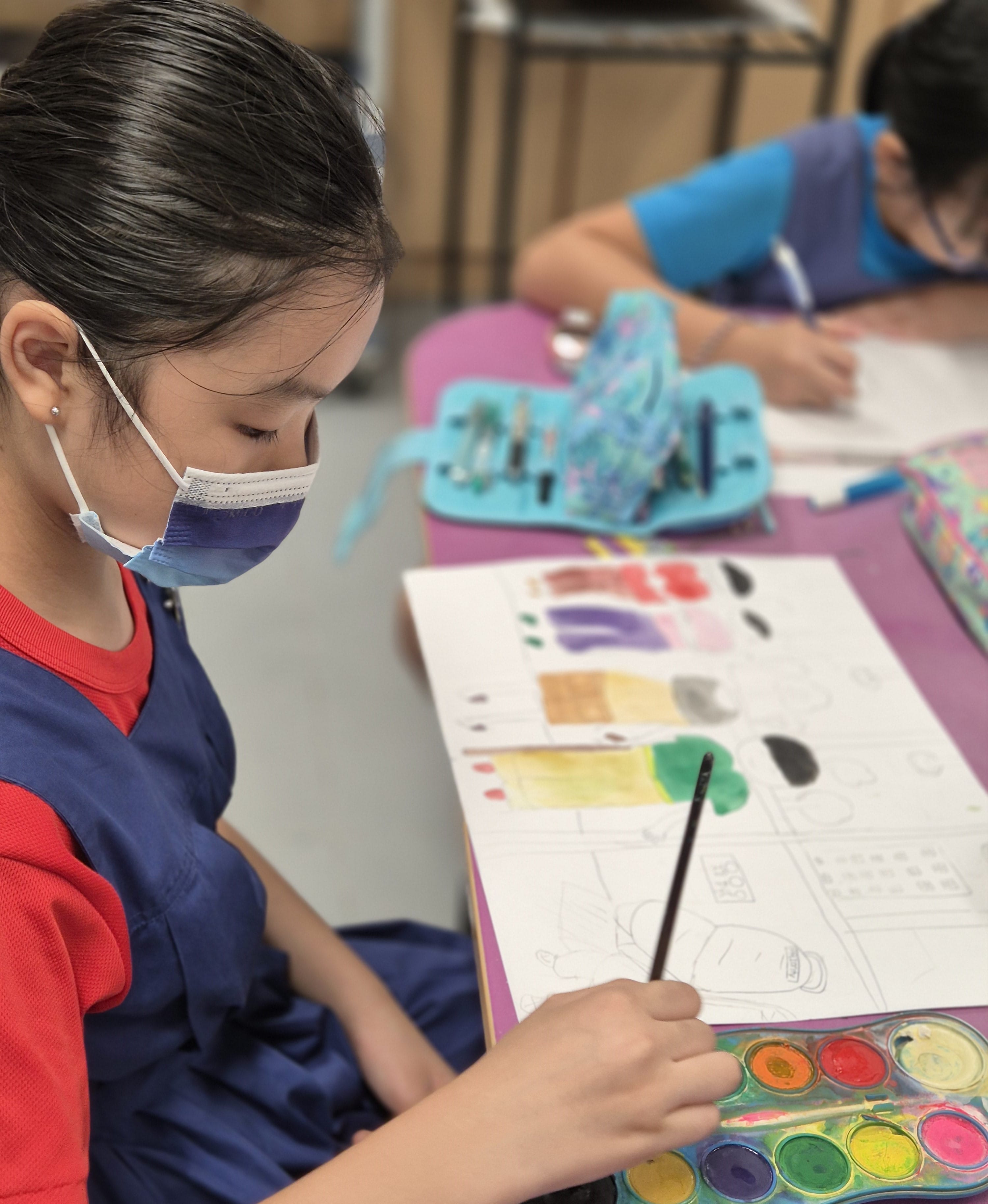 An Art Club member paints colorful watercolor swatches on paper at a desk, with another masked child working in the background.