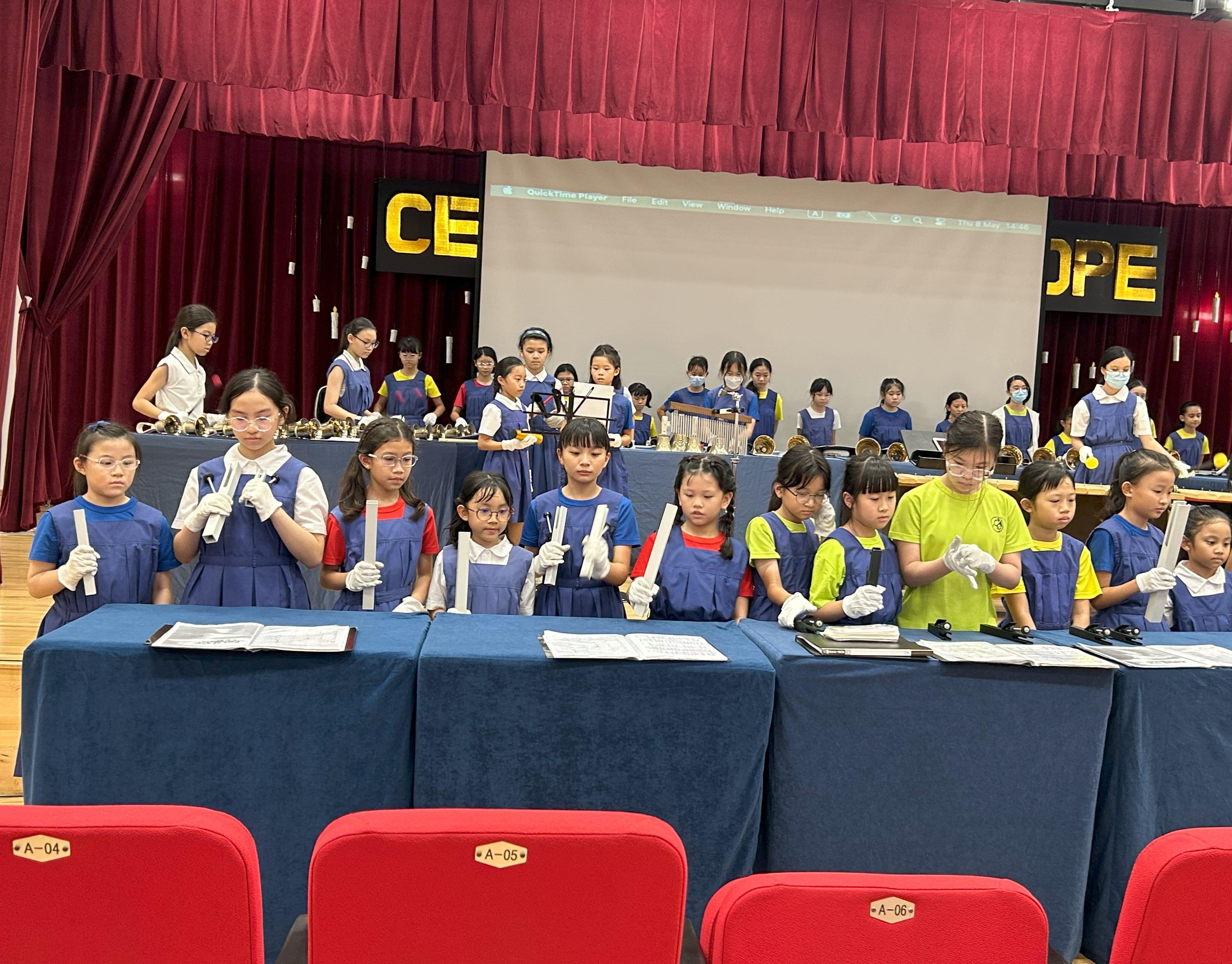 Young girls in blue aprons wearing white gloves hold handbells at blue-draped tables on a school auditorium stage.