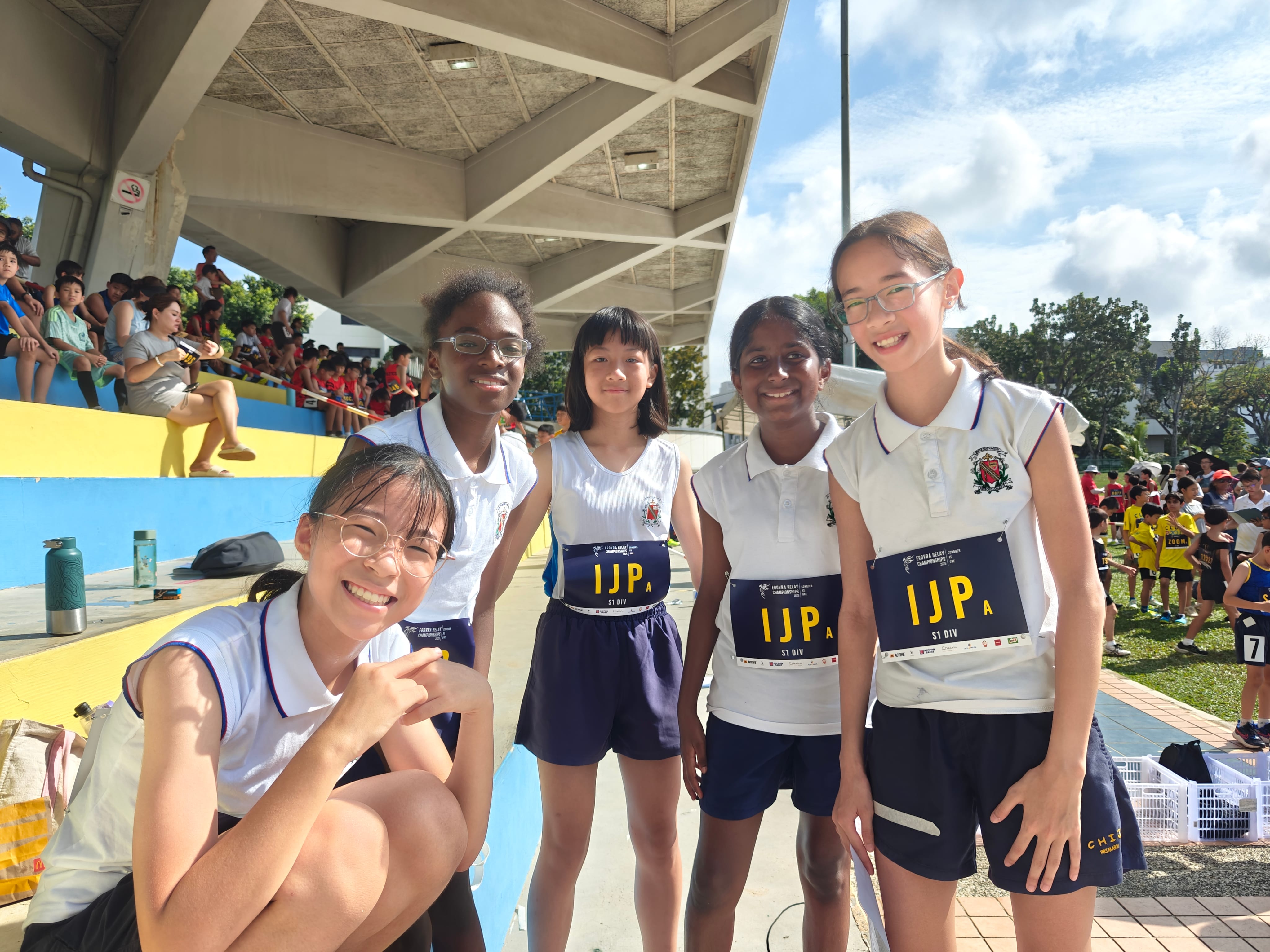 Five girls in white sports uniforms with IJP bibs pose smiling at a track meet beside stadium seats.