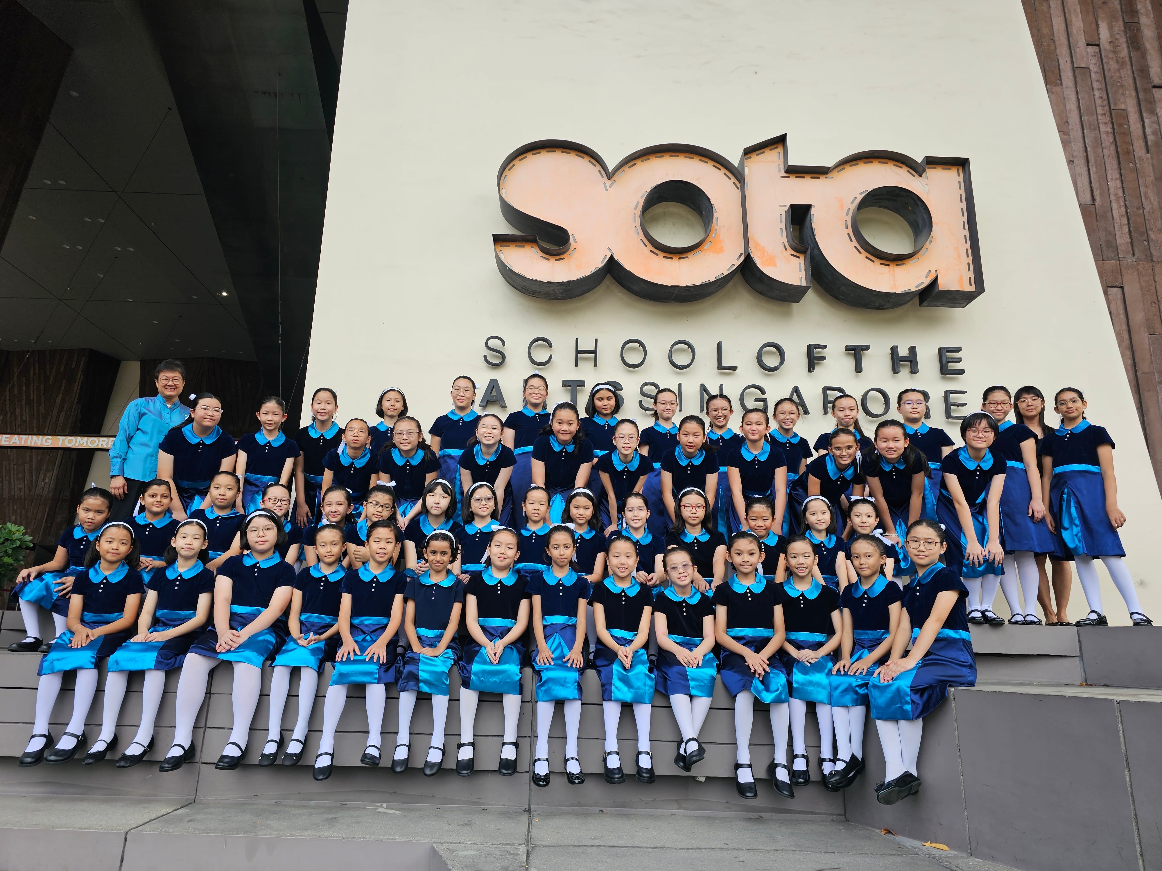A large group of girls in navy and blue uniforms pose in front of the School of the Arts Singapore sign.