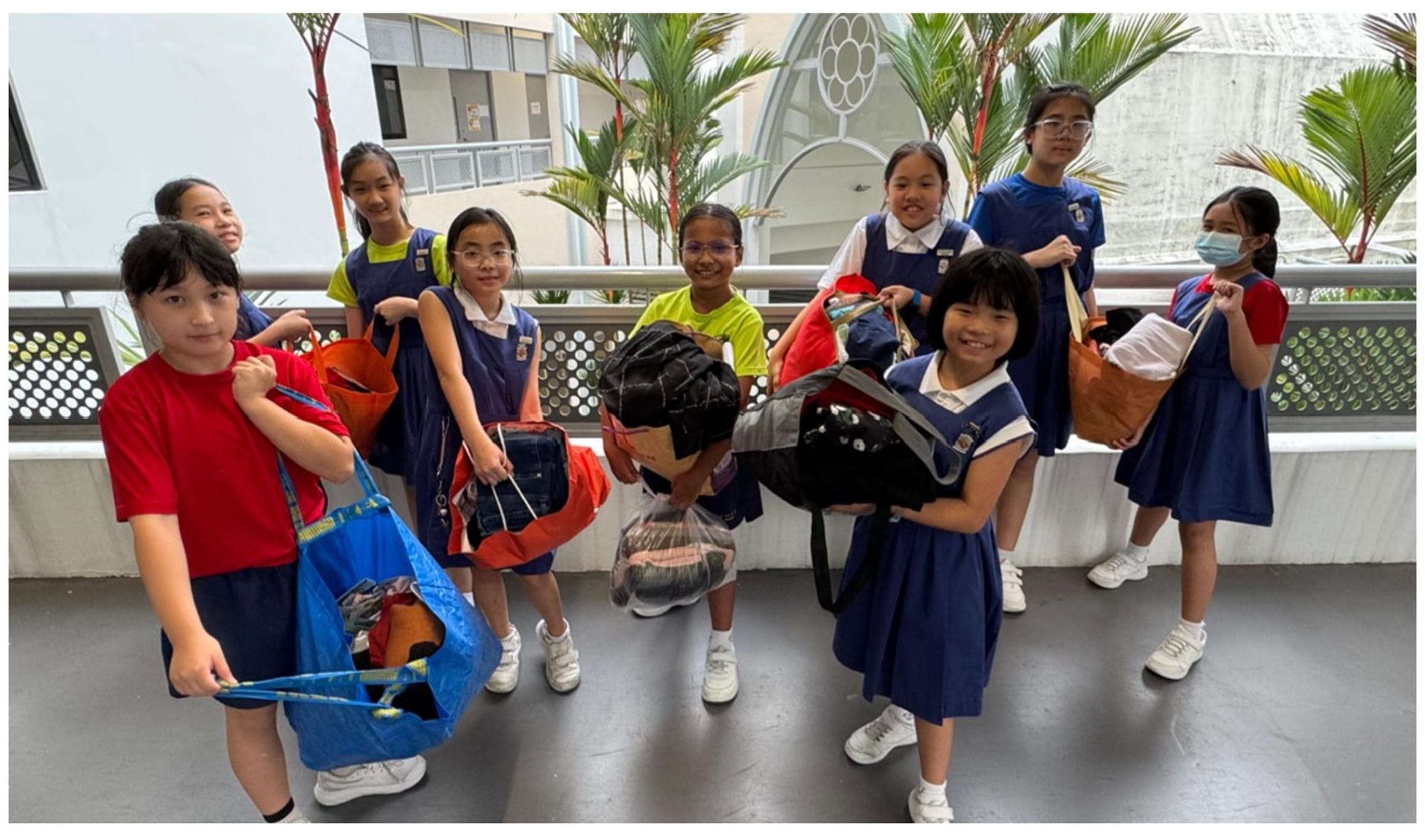 Eight young girls in school uniforms stand on a walkway holding bags filled with donated clothing items.
