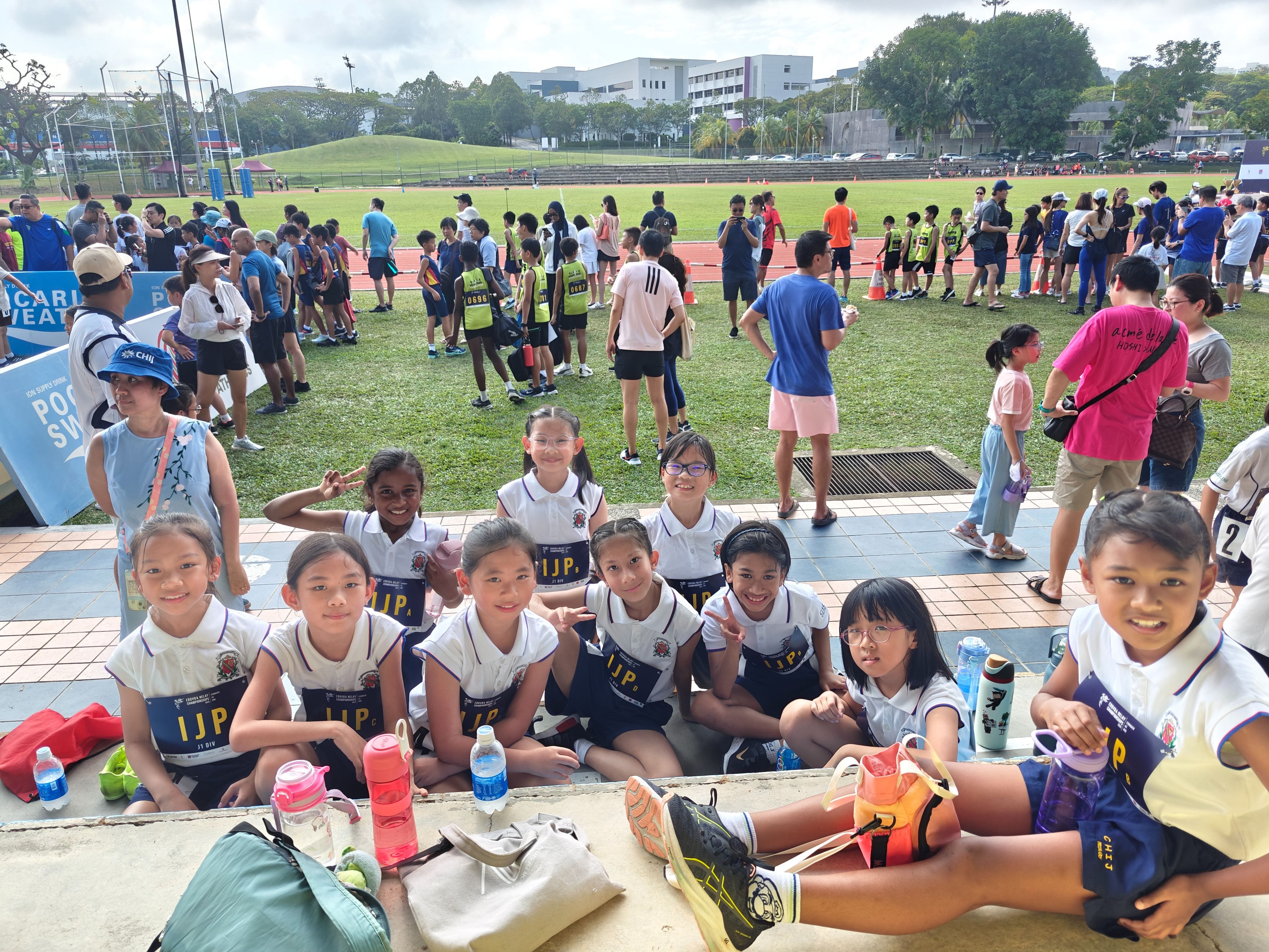 Young students in school uniforms sit together at a crowded outdoor track and field sports event.